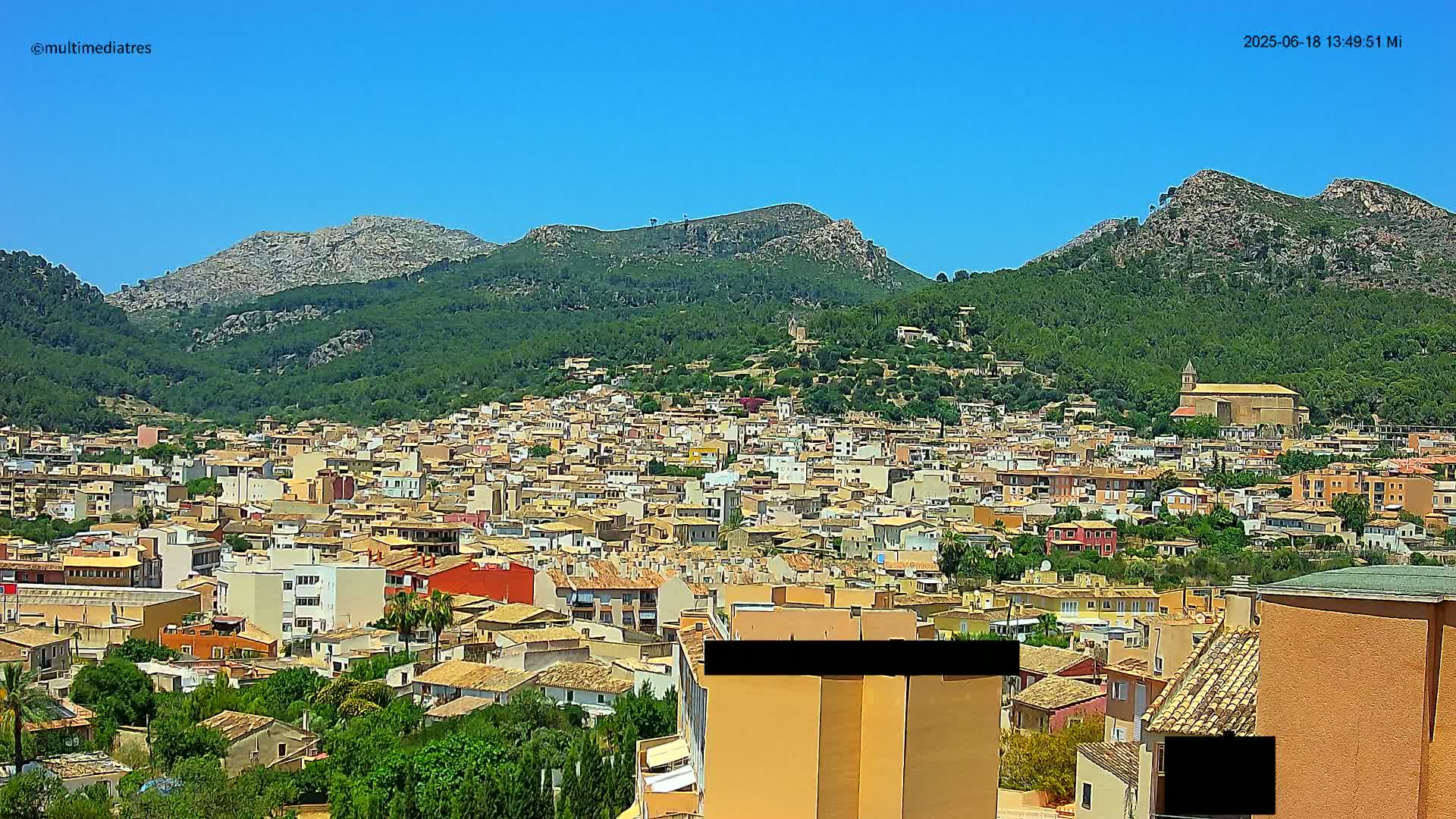 A dense cluster of buildings sits nestled at the base of a large, verdant mountain range under a clear blue sky.