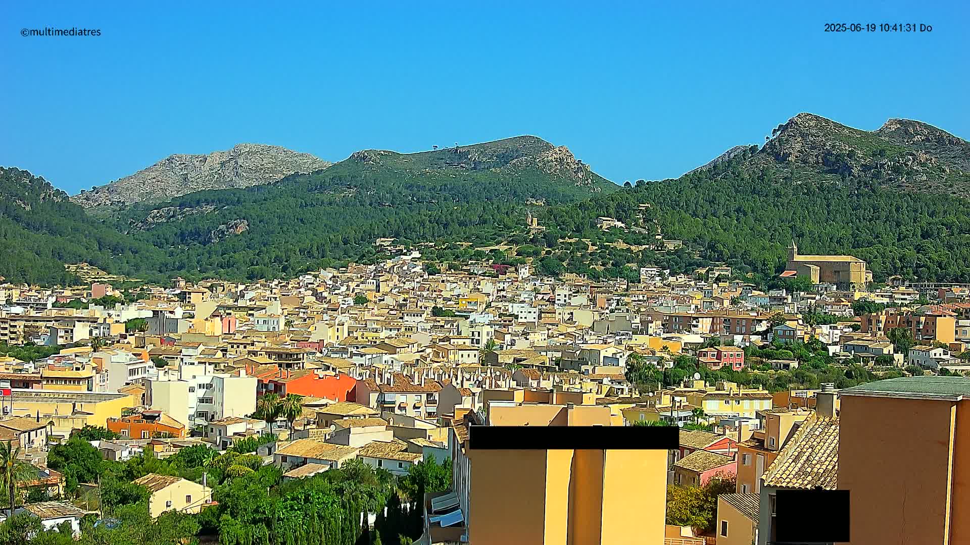 A sprawling town of low-rise buildings nestles in a valley surrounded by lush green hills and rocky mountains under a clear blue sky.