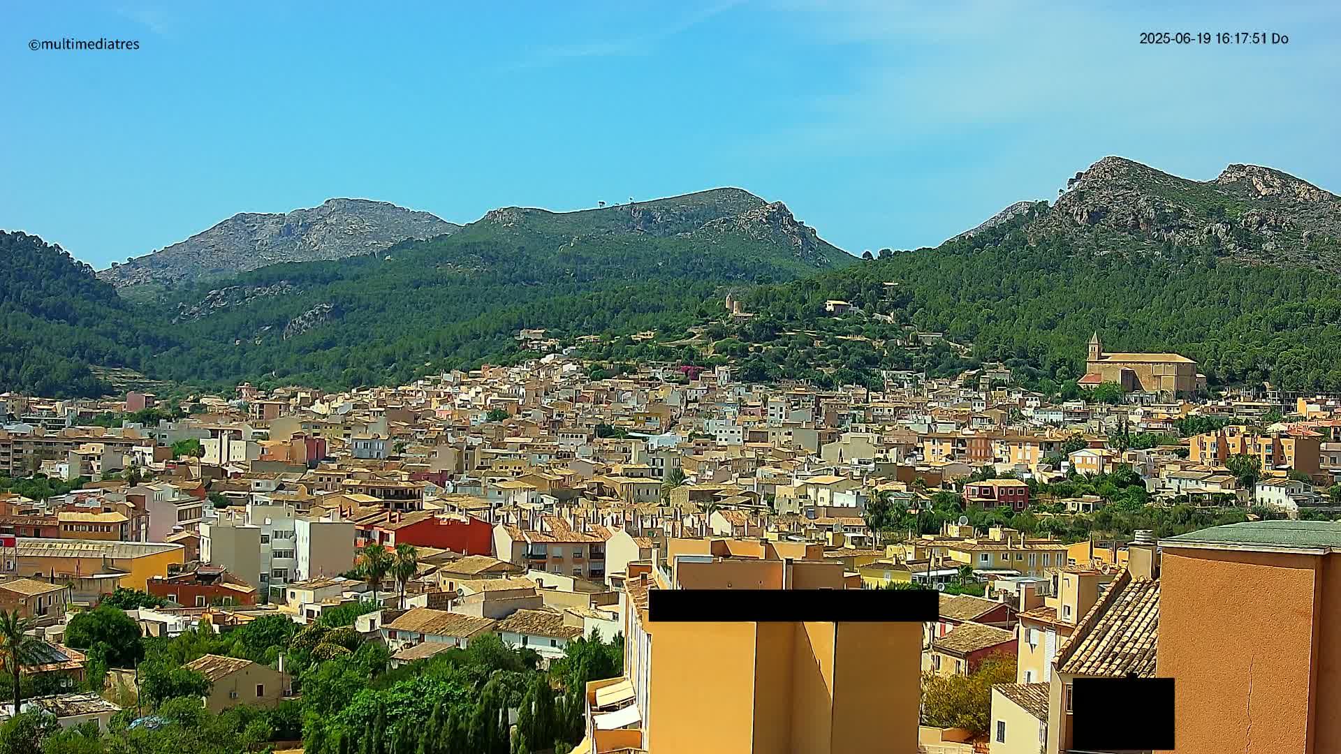 A sprawling town of low-rise buildings nestles at the base of lush green mountains under a clear blue sky.