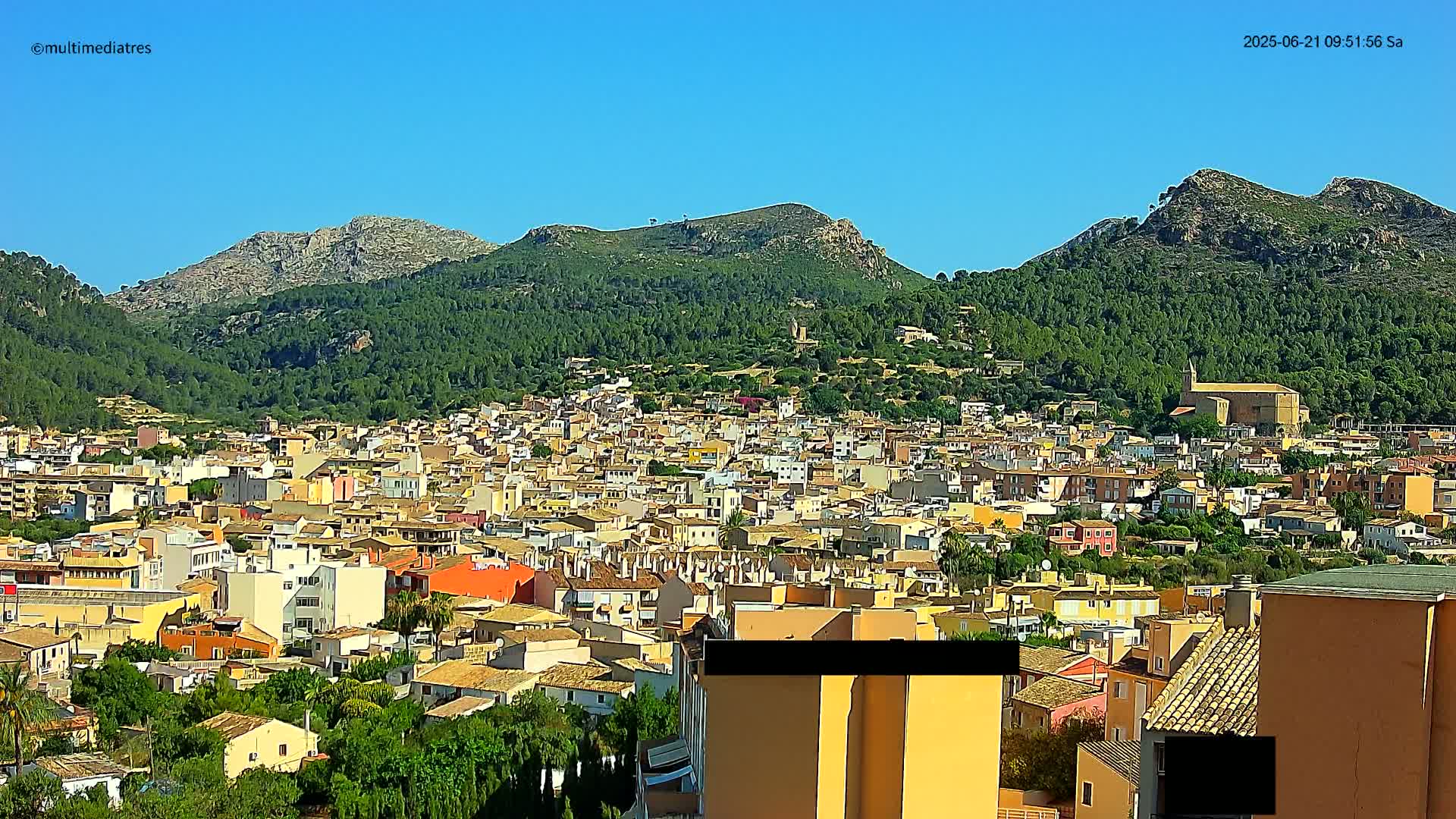 A densely populated town nestled in a valley surrounded by green, mountainous terrain under a clear blue sky.