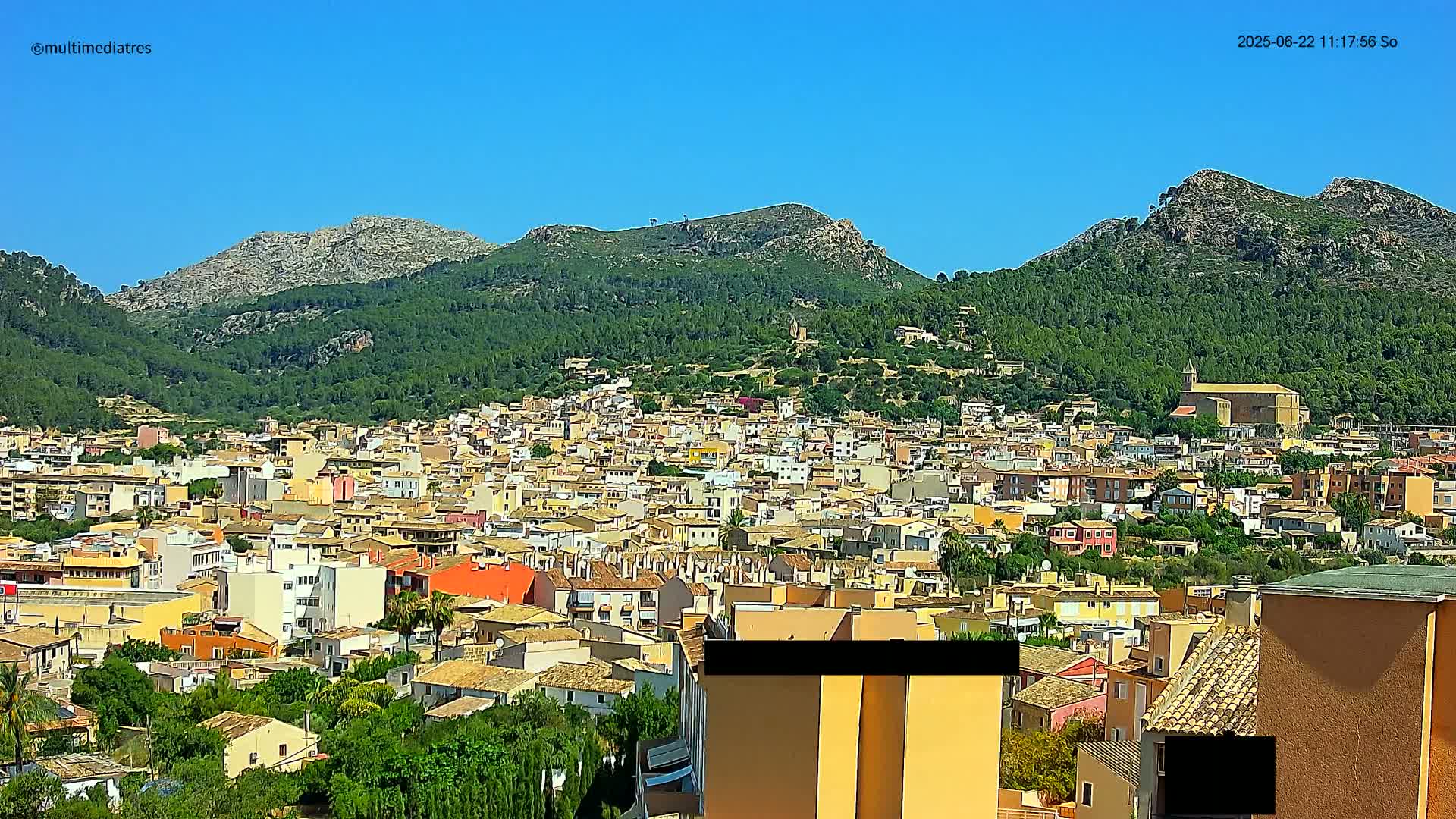 A sprawling town of low-rise buildings nestled among green hills under a clear blue sky.