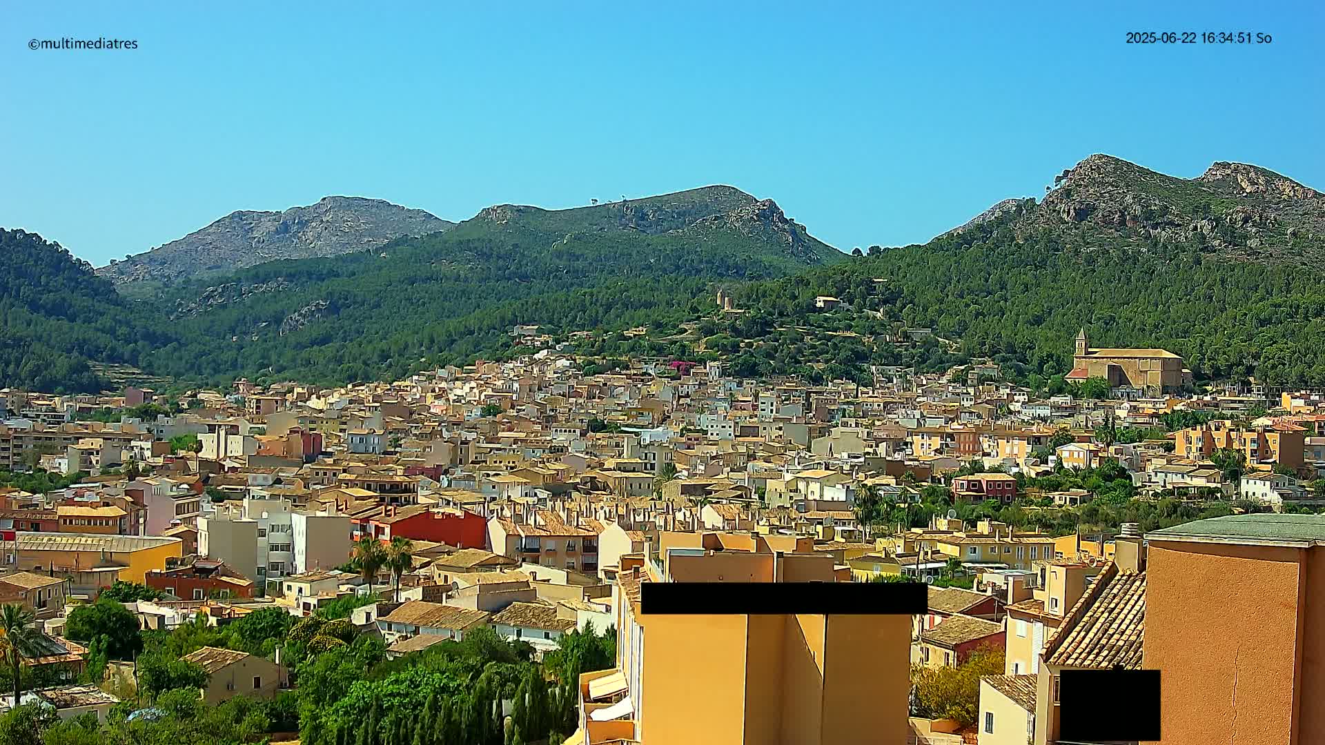 A sprawling town of closely packed, low-rise buildings nestles in a lush green valley, backed by rocky, tree-covered mountains under a clear blue sky.