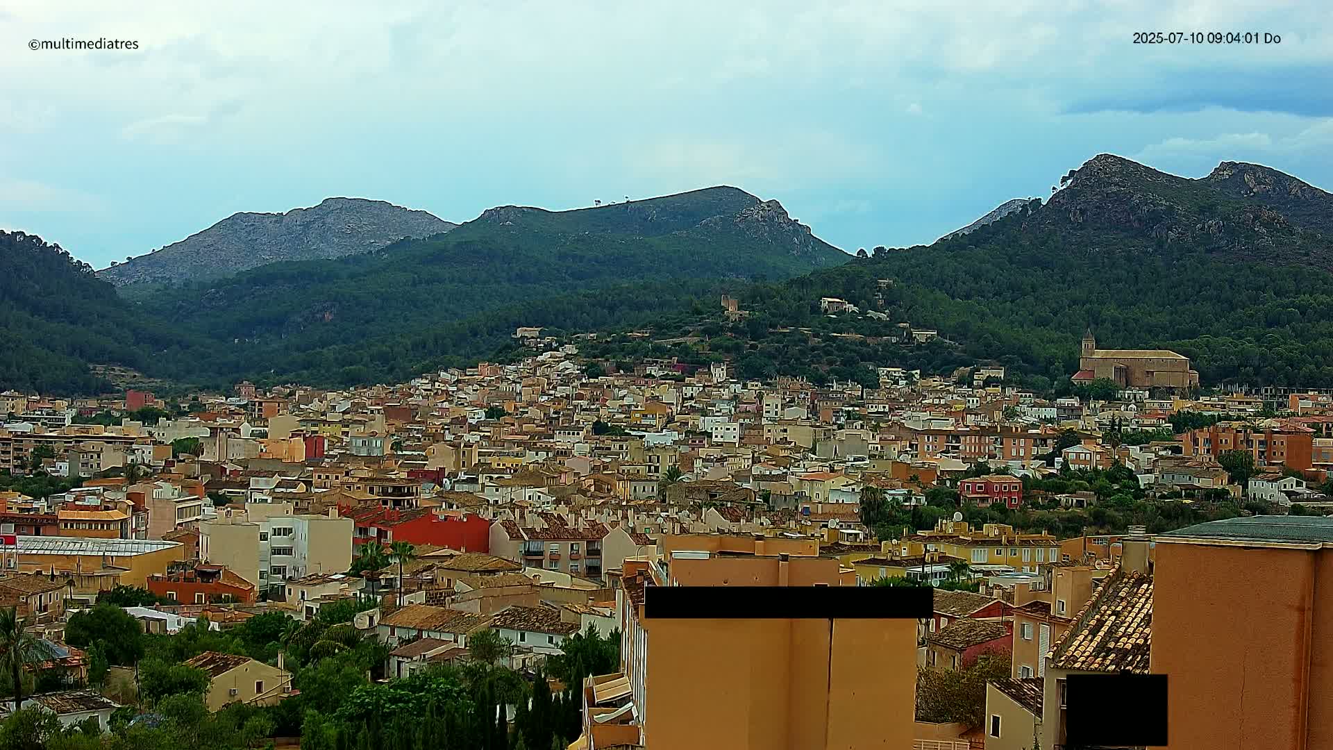 A sprawling town of low-rise buildings nestles in a valley surrounded by lush green hills and mountains under a partly cloudy sky.