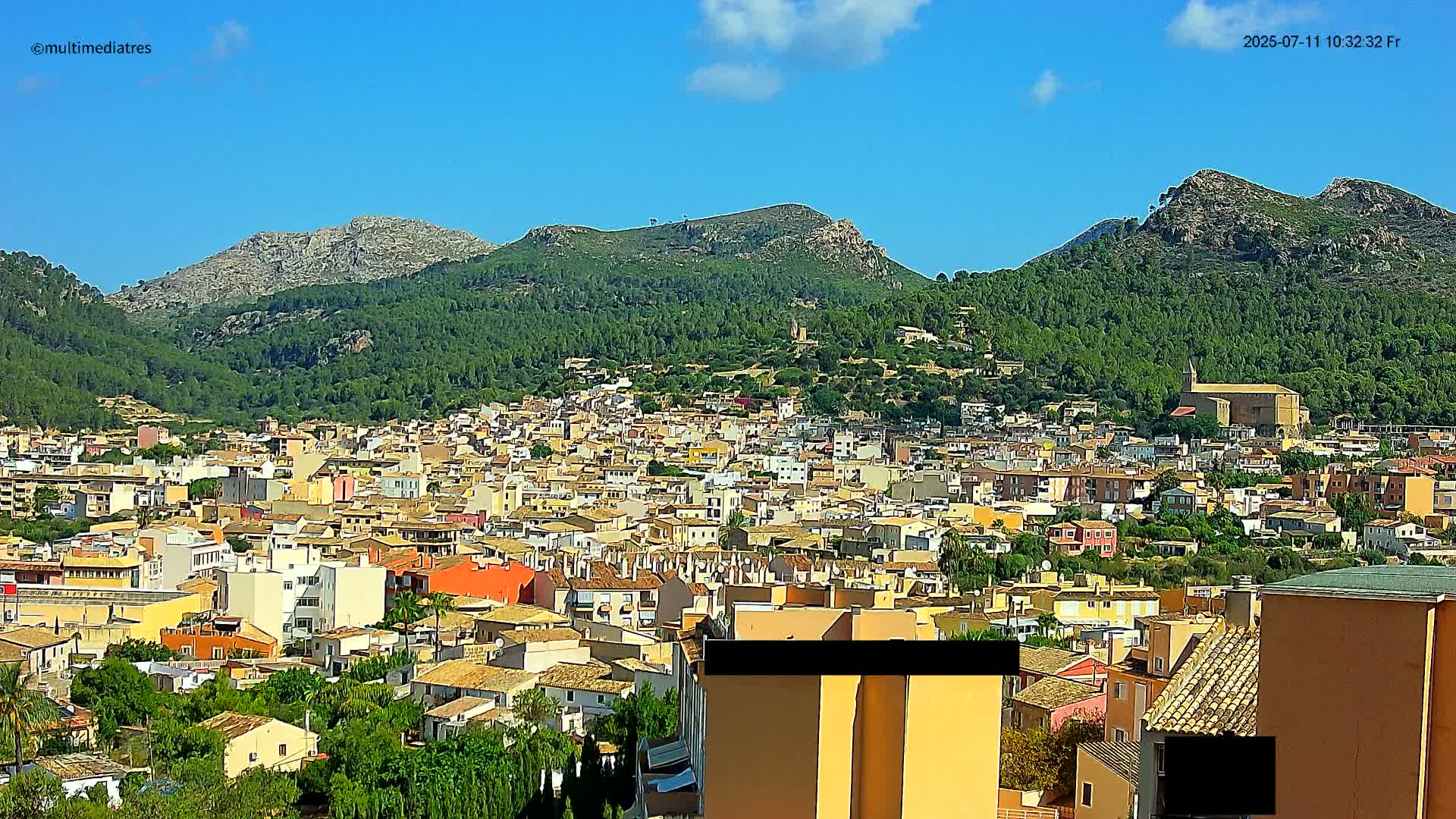 A sprawling town of low-rise buildings nestles at the base of lush green mountains under a clear blue sky.