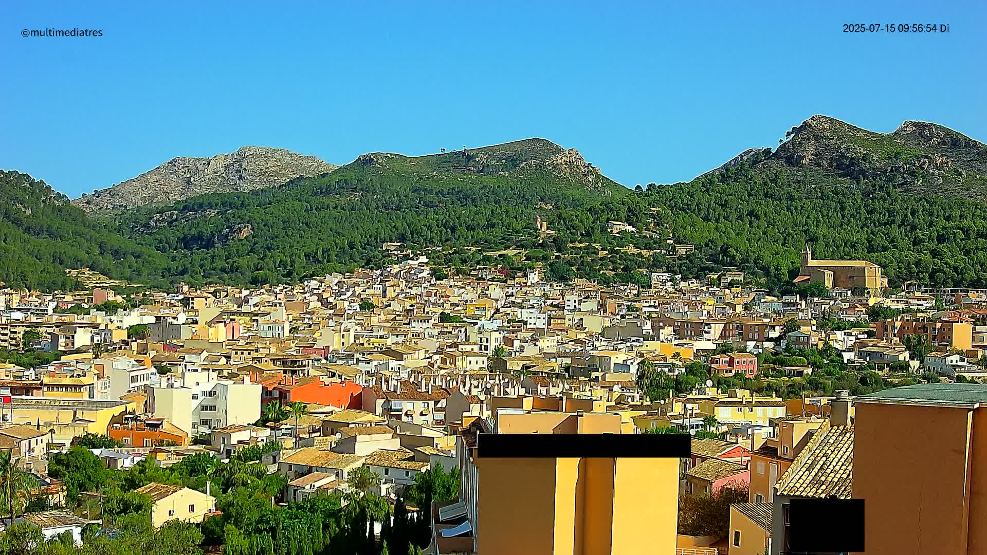 A sprawling town of low-rise buildings nestles against a backdrop of lush green hills and rocky mountains under a clear blue sky.