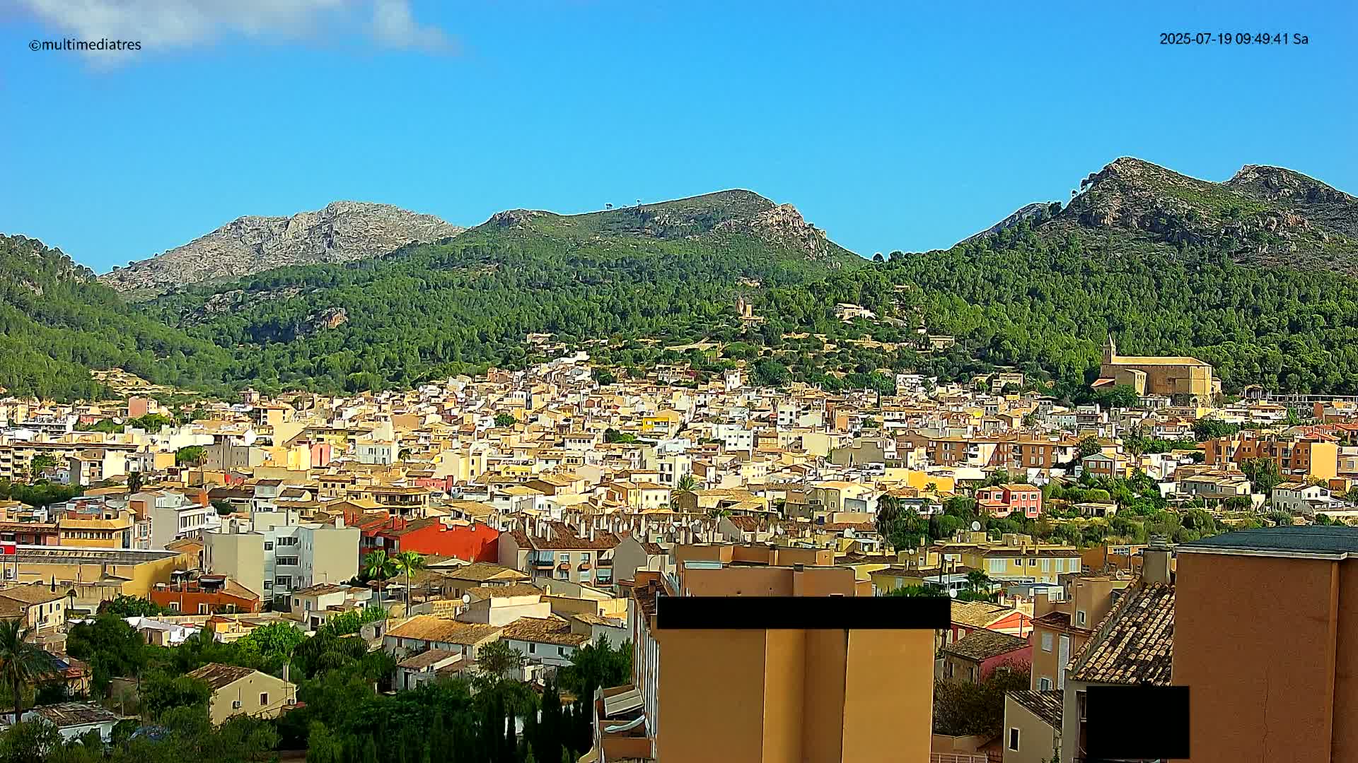 A densely packed town of tan and orange buildings nestled at the base of lush green mountains under a clear blue sky.