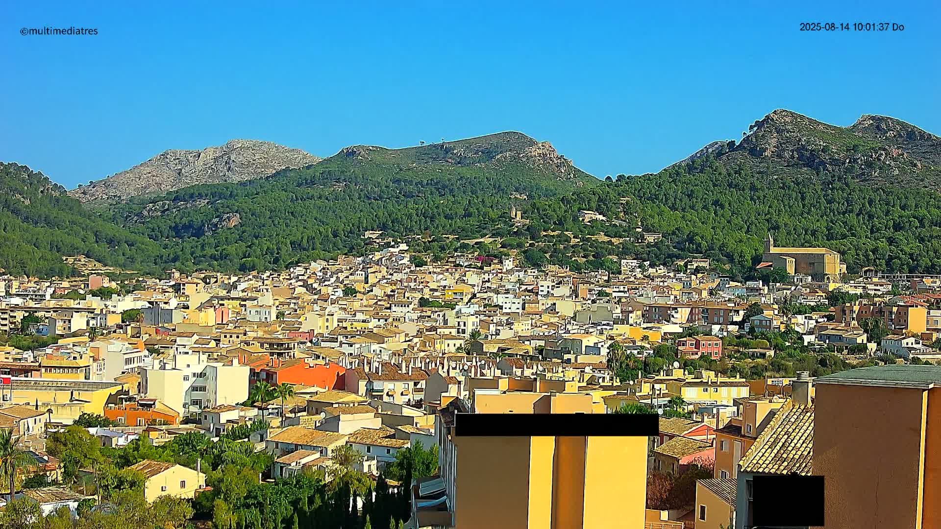 A sprawling town of low-rise buildings nestled at the base of verdant, rocky mountains under a clear blue sky.