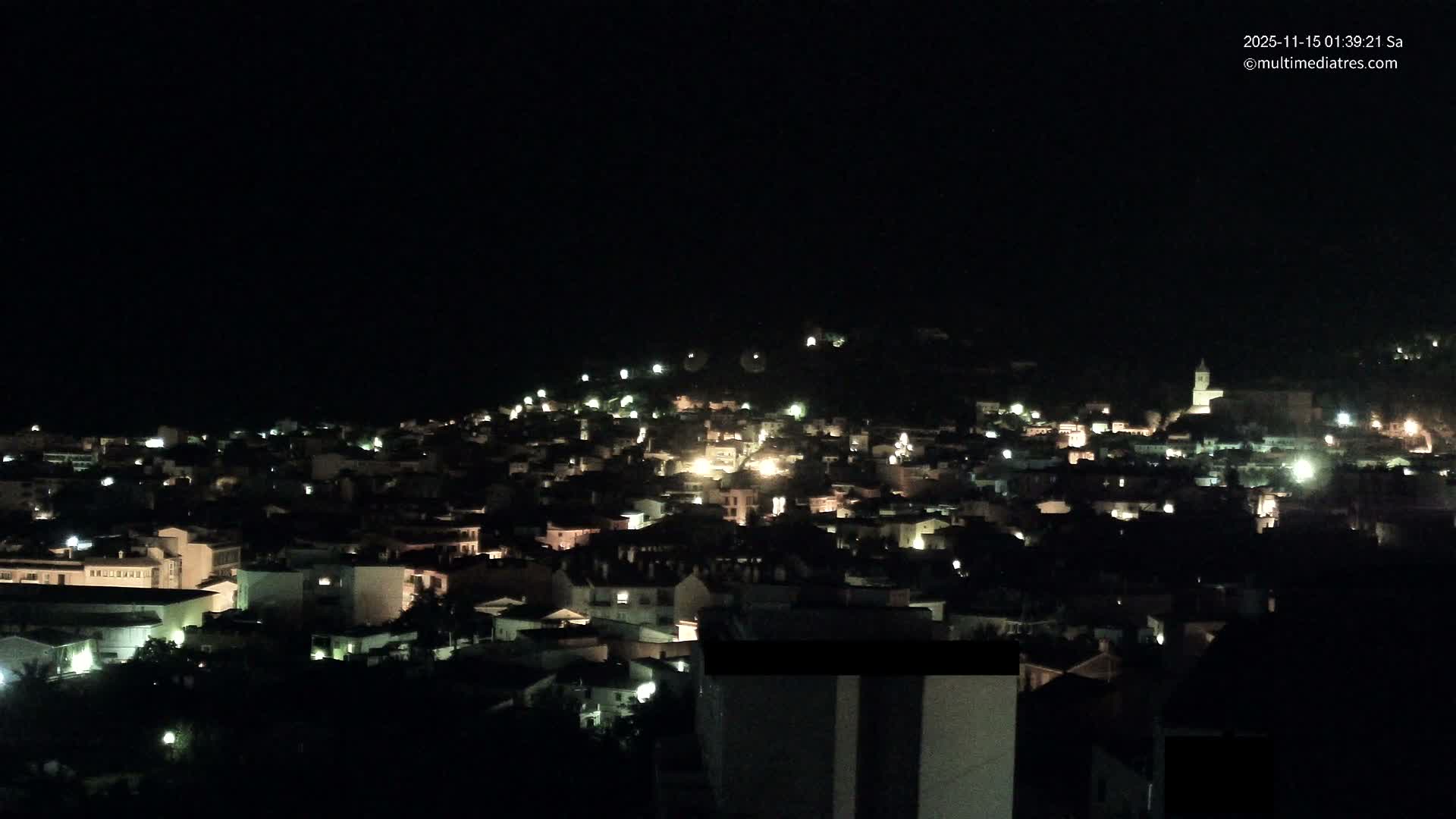Under a clear night sky, a hillside town is brightly illuminated by numerous street and building lights, featuring a prominent church steeple among its structures.