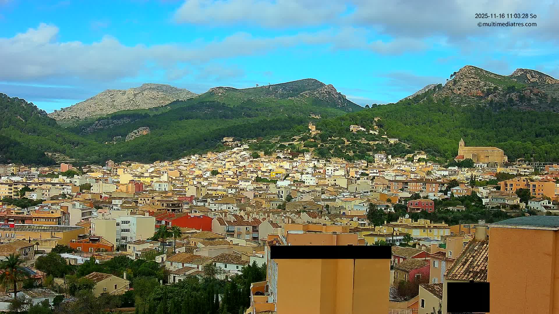 A vibrant town with pastel and terracotta buildings fills a valley, nestled amongst lush green mountains under a partly cloudy blue sky, with a large church prominent on the right.