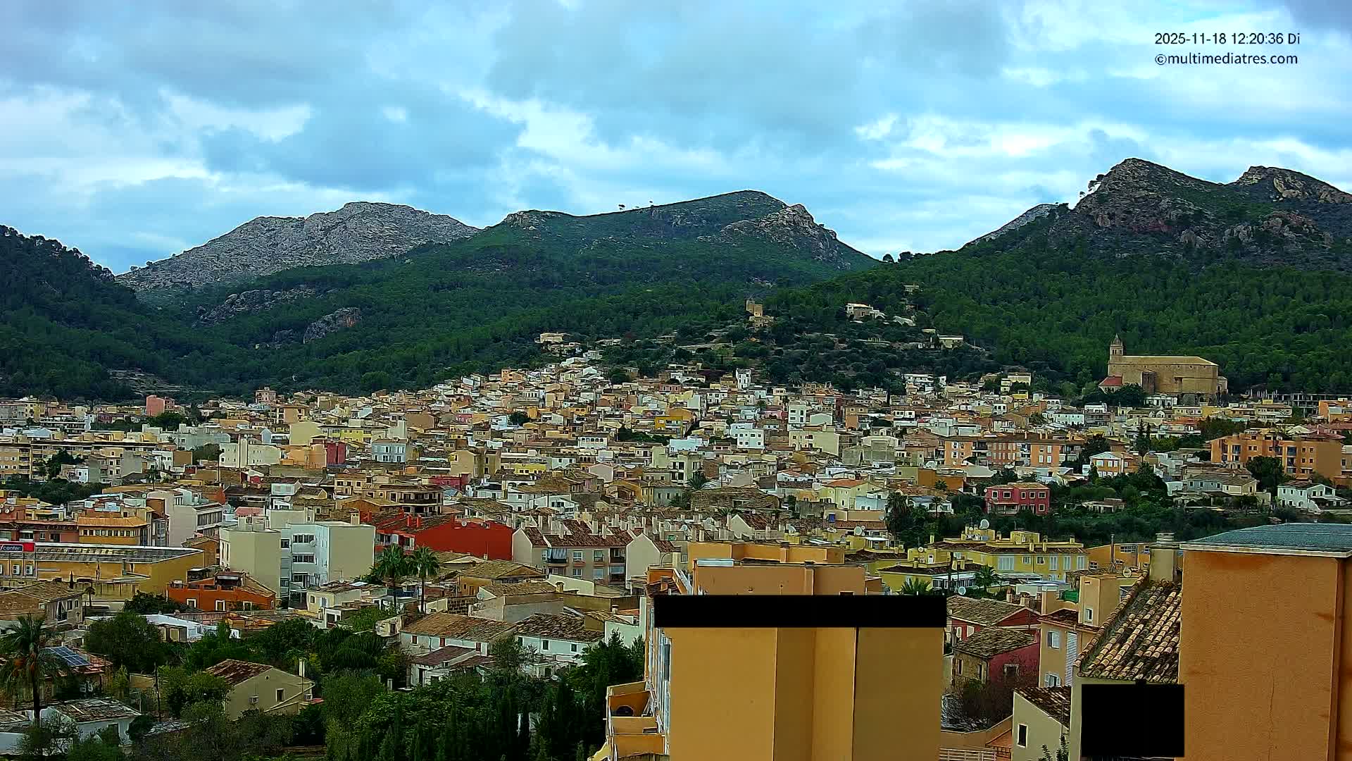 A vibrant town with pastel and terracotta buildings fills a valley, nestled amongst lush green mountains under a partly cloudy blue sky, with a large church prominent on the right.
