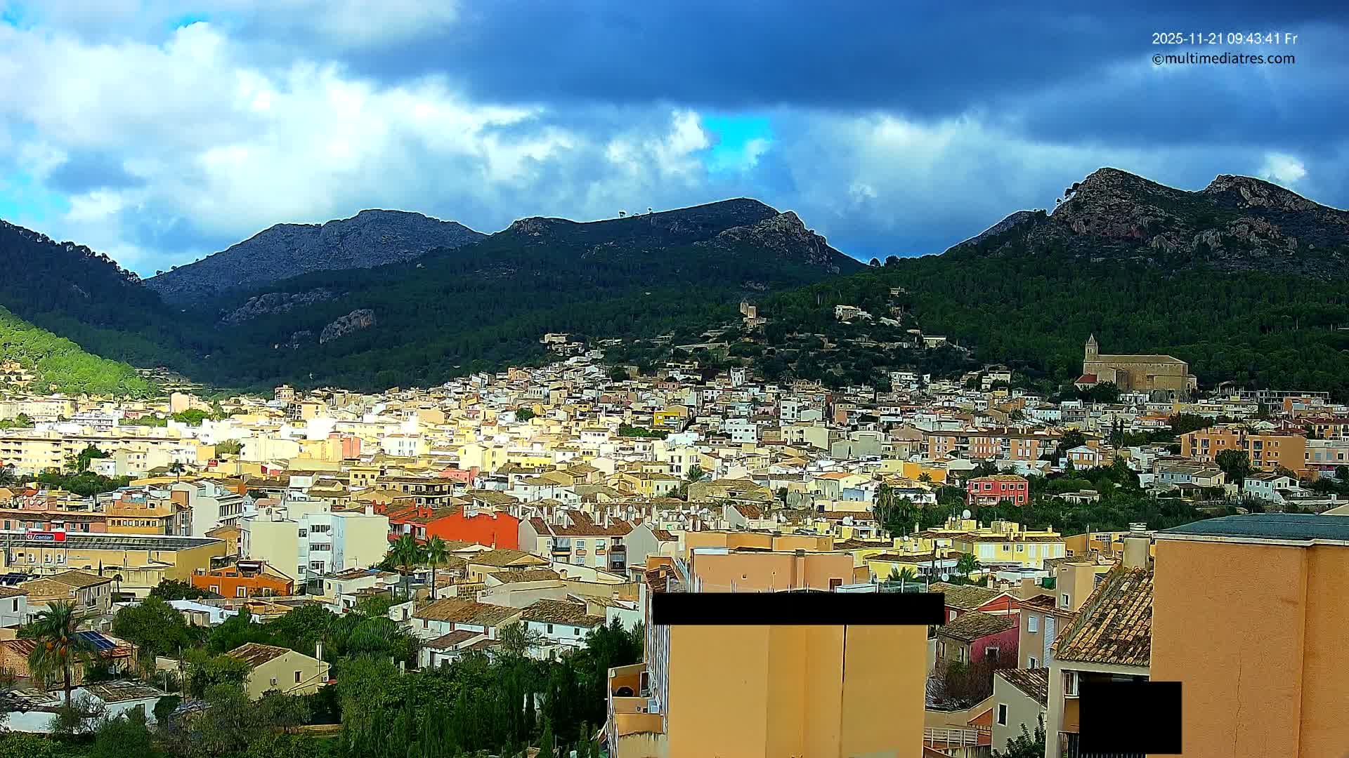 A sprawling hillside town with a mix of colorful and light-toned buildings and terracotta roofs is nestled between large, tree-covered mountains, under a partly cloudy sky with sun breaking through.