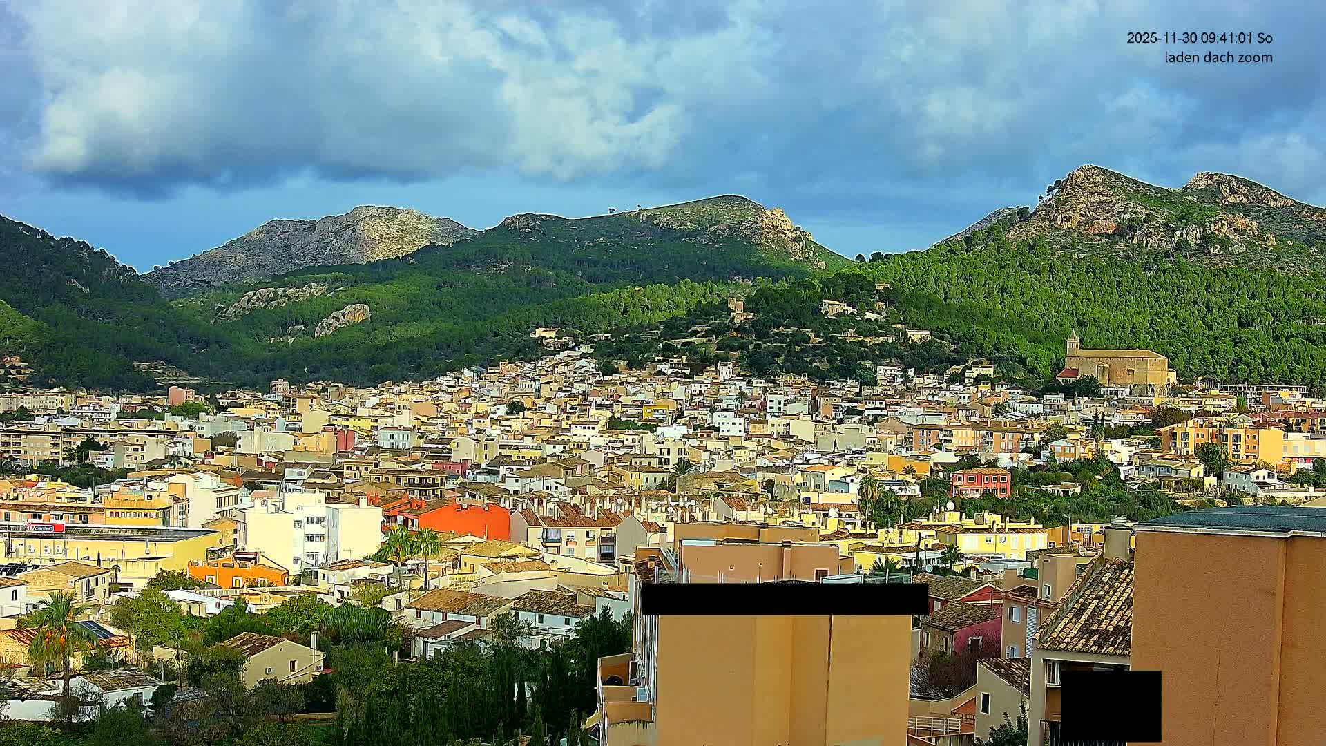 An expansive view reveals a colorful town nestled at the base of lush, forested mountains under a partly cloudy sky.