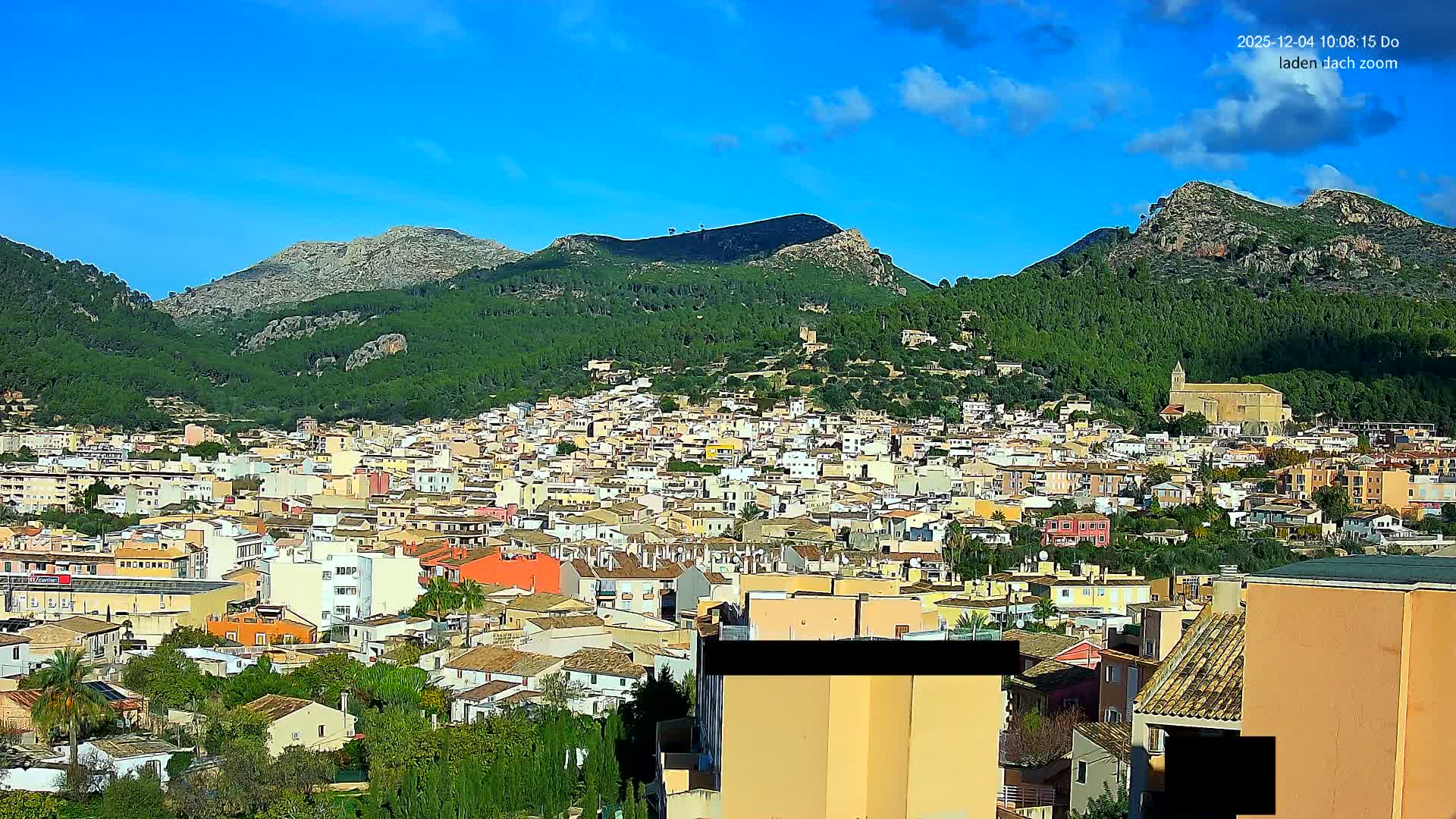 A vibrant town with colorful buildings and a prominent church is nestled among lush green, tree-covered mountains under a bright blue sky with scattered white clouds.