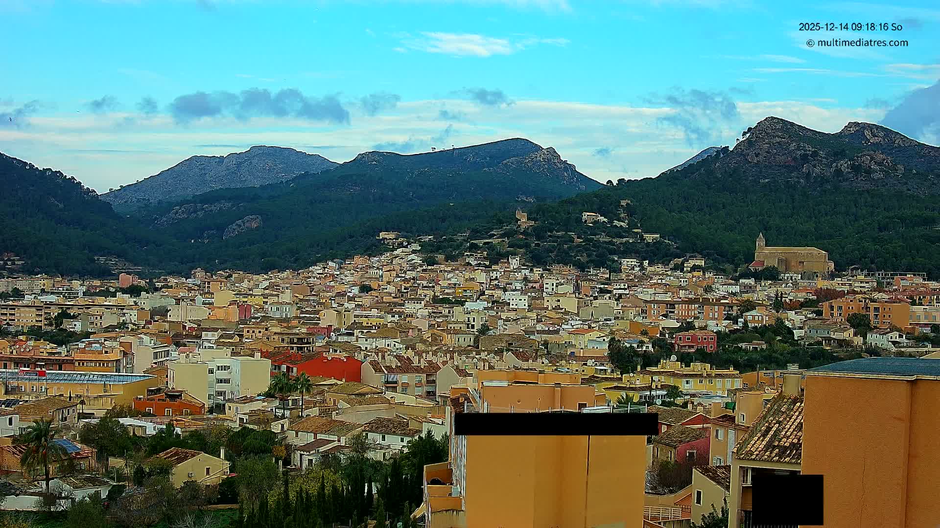 A vibrant town with colorful buildings and a prominent church is nestled among lush green, tree-covered mountains under a bright blue sky with scattered white clouds.
