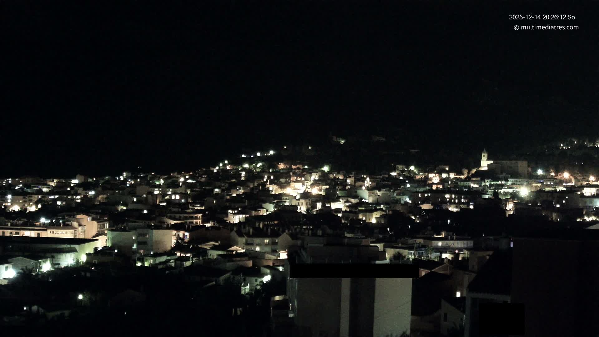 A vibrant town with colorful buildings and a prominent church is nestled among lush green, tree-covered mountains under a bright blue sky with scattered white clouds.