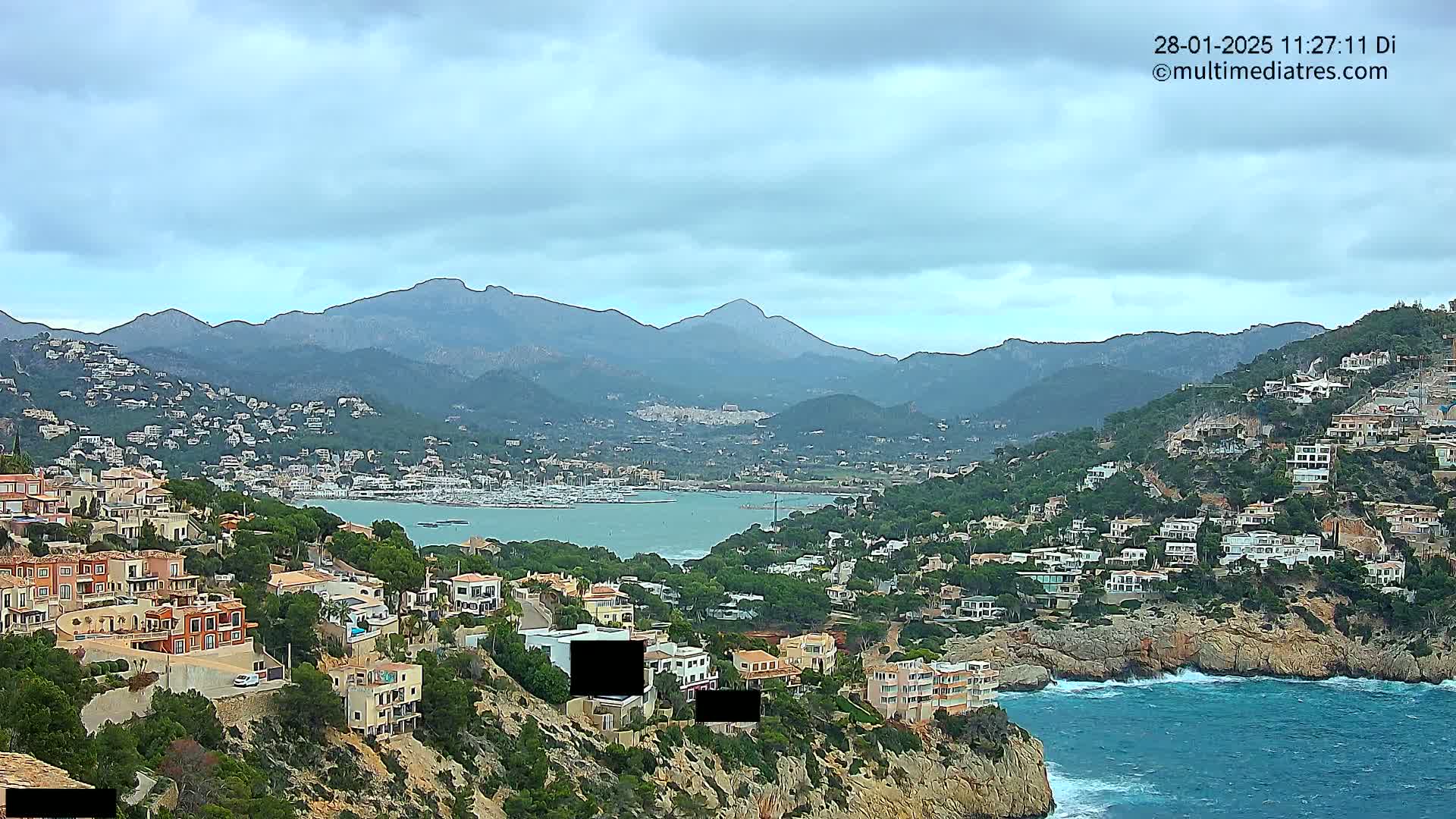 A coastal town nestled in a mountainous region under a cloudy sky, with a calm bay and a marina visible in the distance.