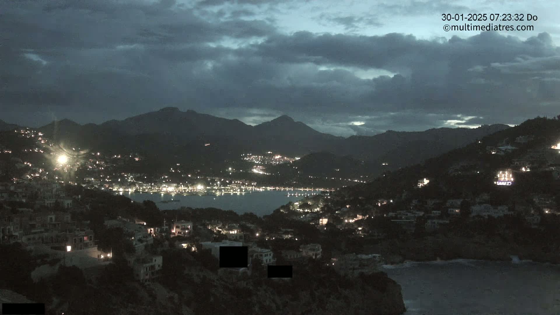 A nighttime view of a coastal town nestled in a mountainous region under a cloudy sky.