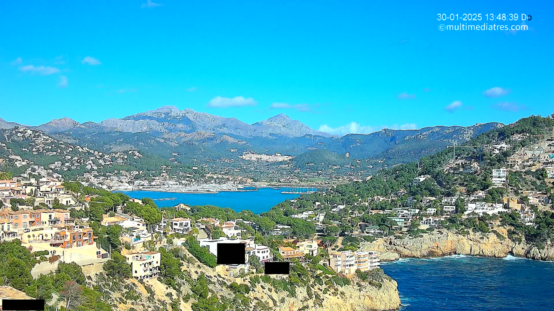 A coastal town nestled on a hillside overlooking a calm bay, with mountains in the background under a clear blue sky.