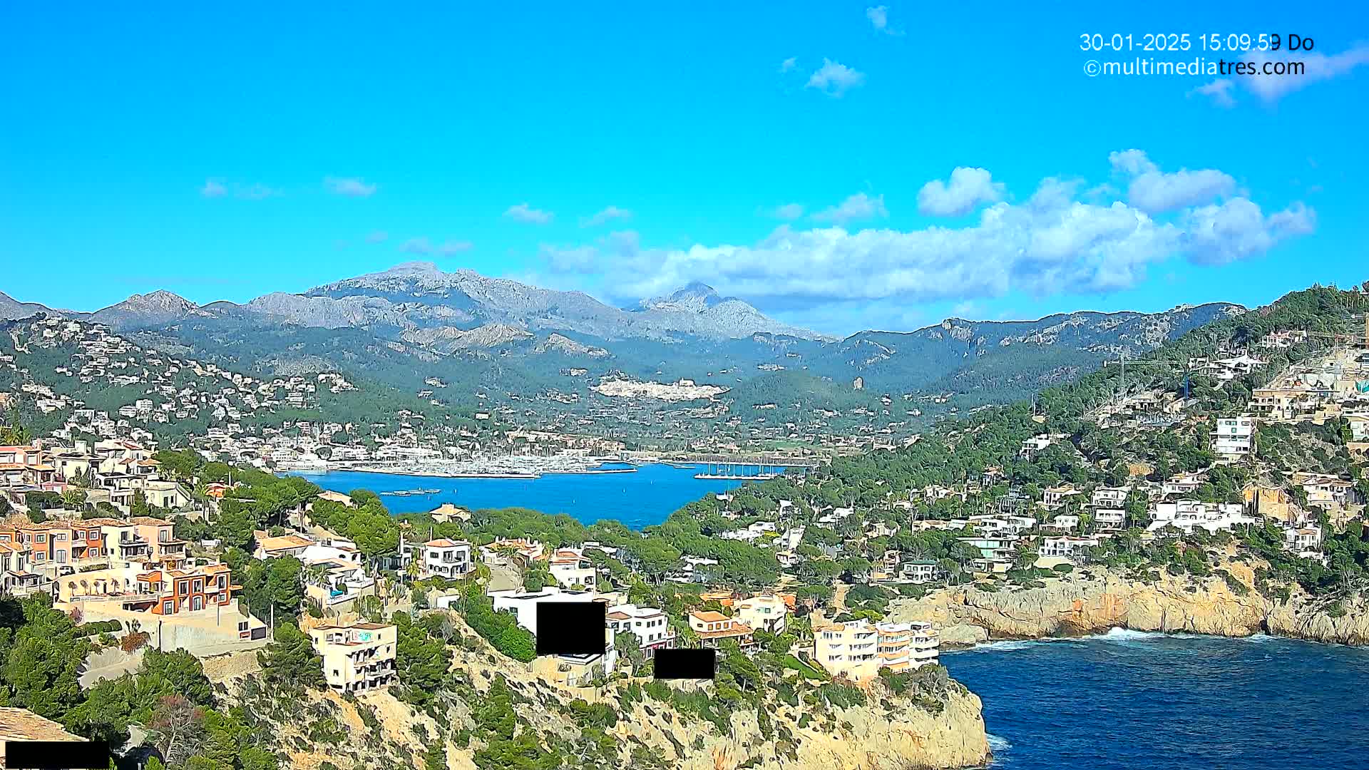 A coastal town nestled in a mountainous region under a mostly sunny sky, with a calm bay filled with boats visible in the distance.