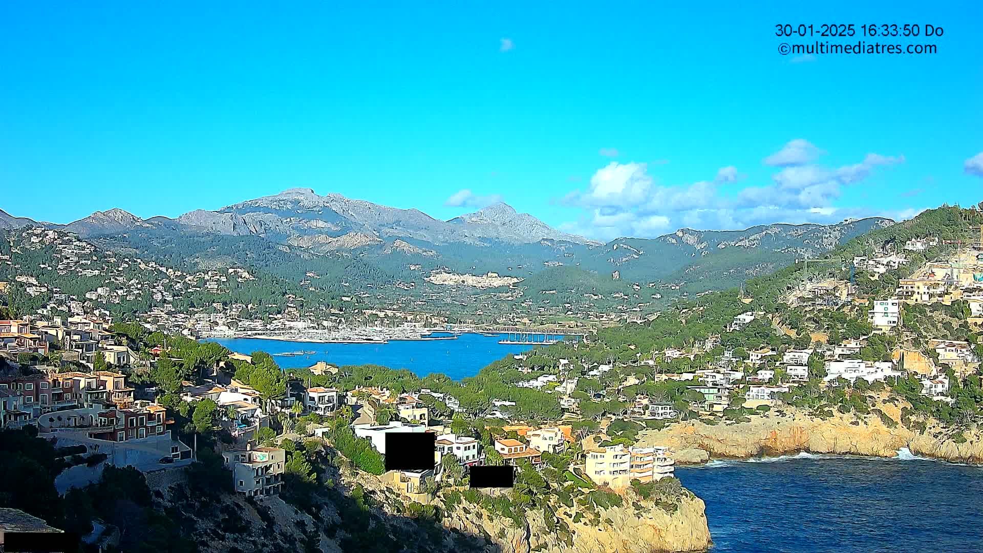 A coastal town nestled against mountains under a clear blue sky with a few scattered clouds, overlooks a calm bay filled with boats.