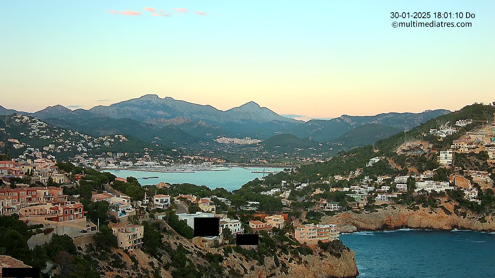 A coastal town nestled in a mountainous region, overlooking a calm bay filled with boats, under a soft, twilight sky.