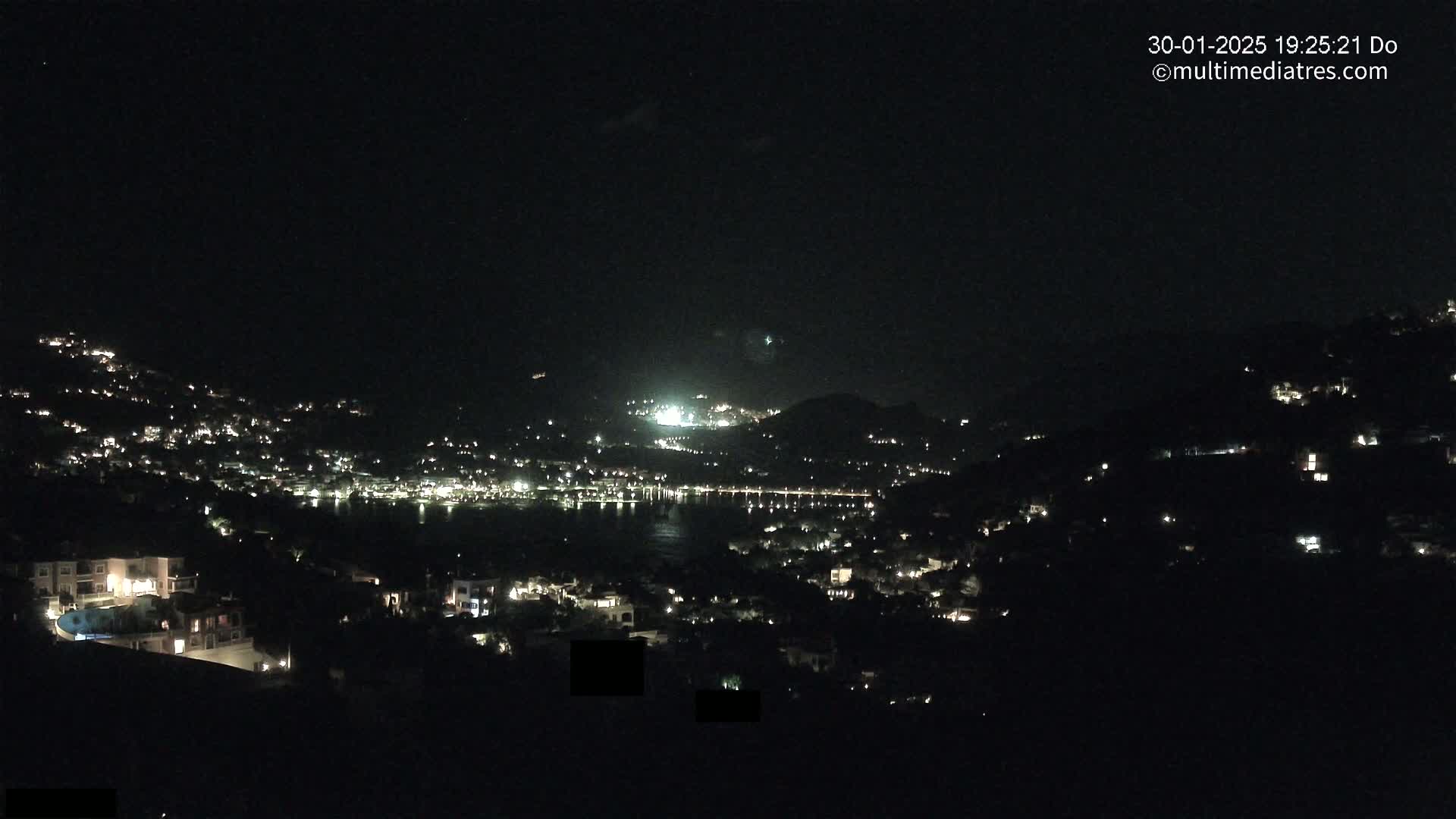 A nighttime view of a coastal town nestled on hillsides, illuminated by scattered lights, overlooking a dark body of water under a clear sky.