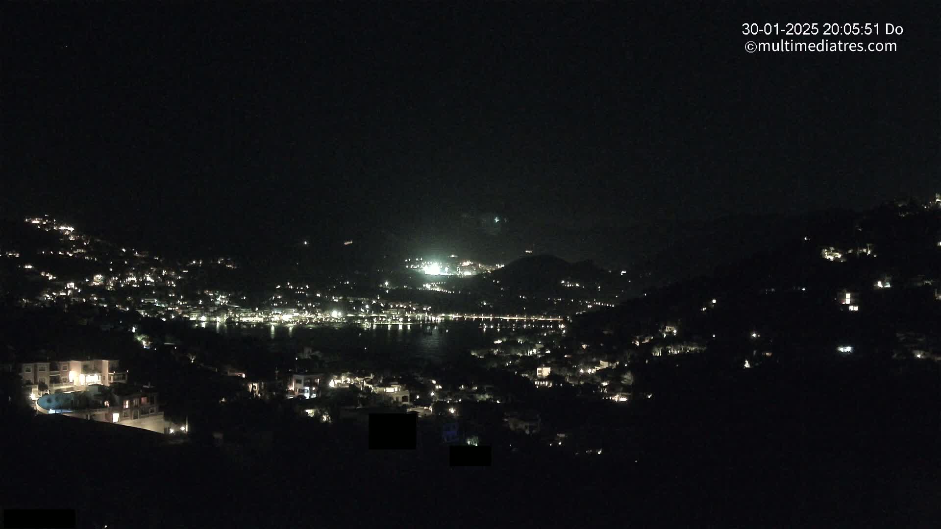 A nighttime view of a coastal town nestled in hills, with scattered lights illuminating the landscape under a clear, dark sky.