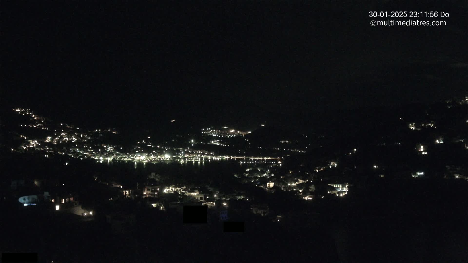 A nighttime view of a town nestled on hillsides surrounding a dark body of water, under a clear, dark sky.