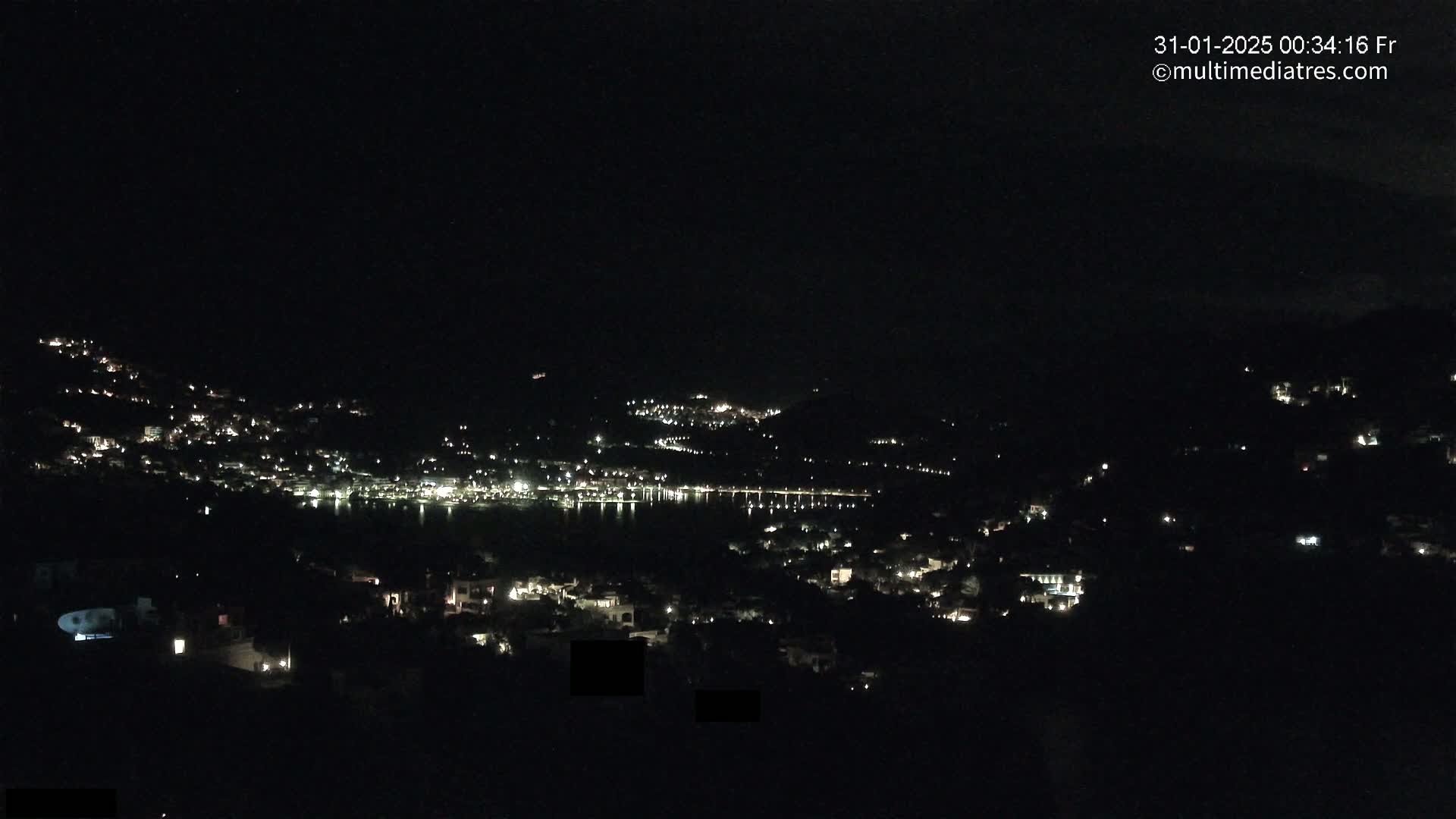 A nighttime view shows a town's lights scattered across hillsides and along a dark body of water.