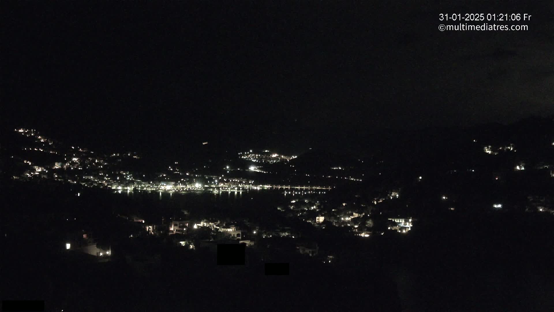 A nighttime view of a coastal town illuminated by scattered lights, with a dark body of water in the middle ground.