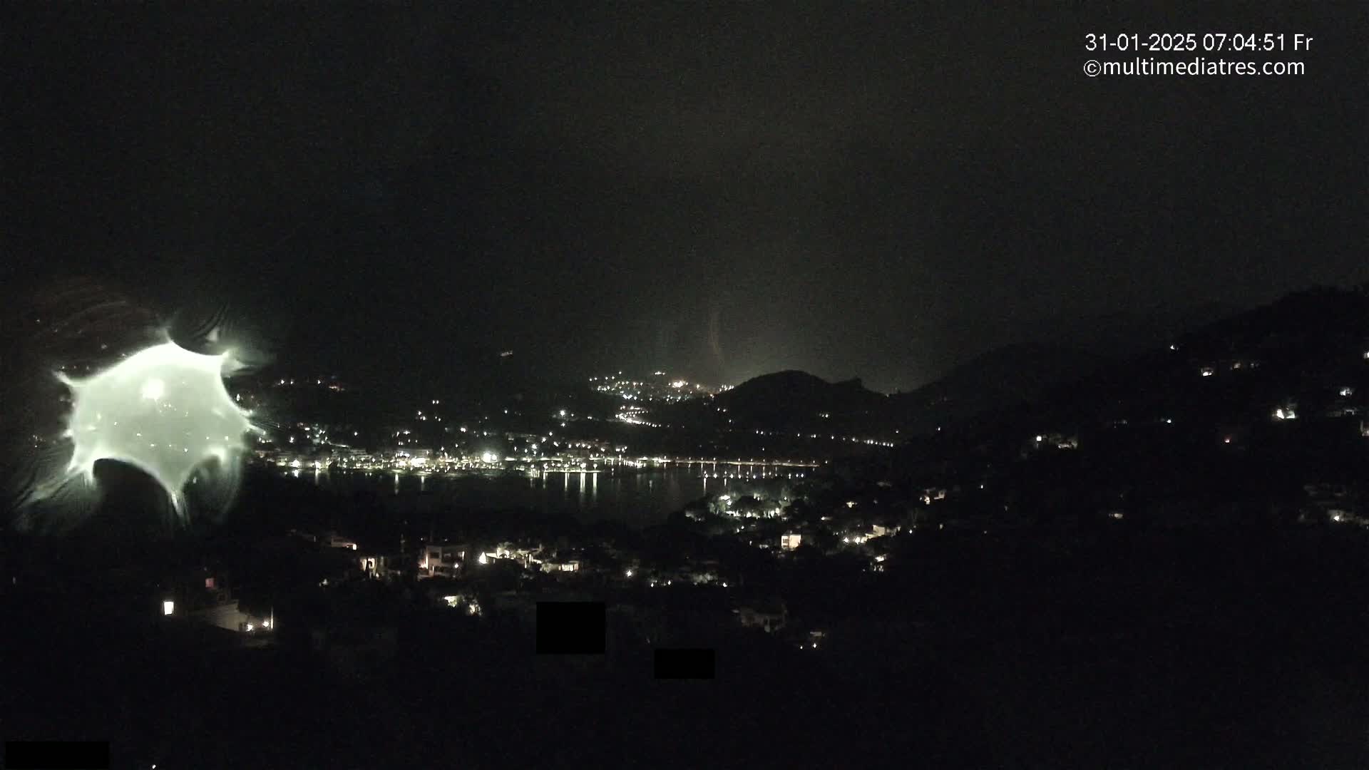 A nighttime view of a coastal town with scattered lights, partially obscured by a blurry, light-colored distortion in the foreground, under a dark, possibly stormy sky.