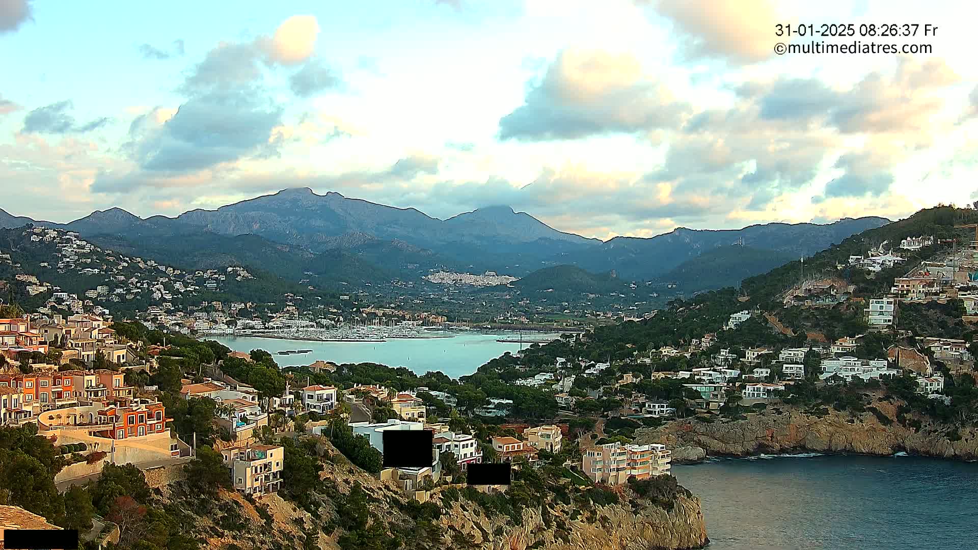 A coastal town nestled in a hillside, overlooking a calm bay filled with boats, with mountains in the background under a partly cloudy sky.