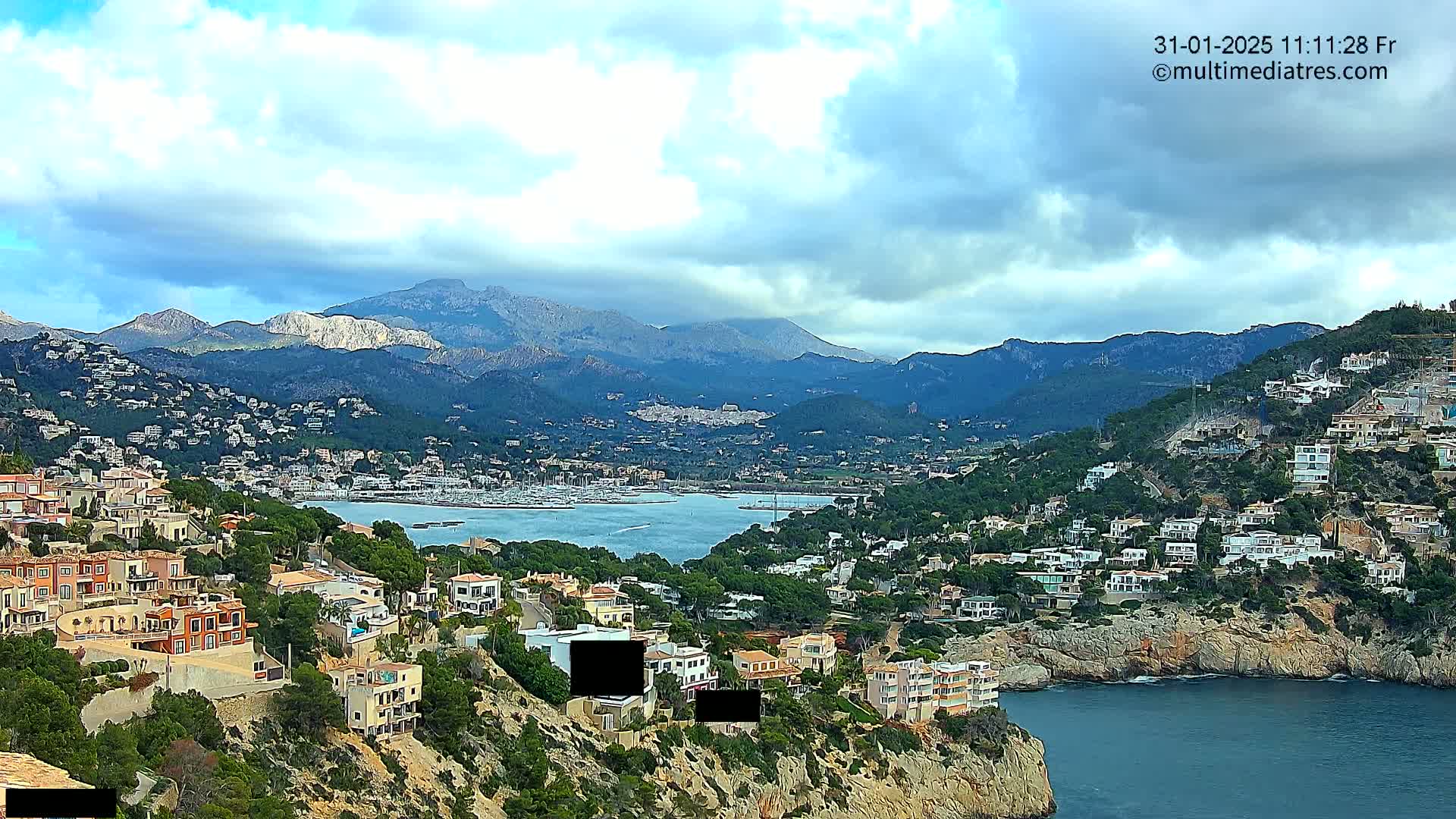 A coastal town nestled on a hillside overlooking a calm bay, with mountains in the background under a partly cloudy sky.