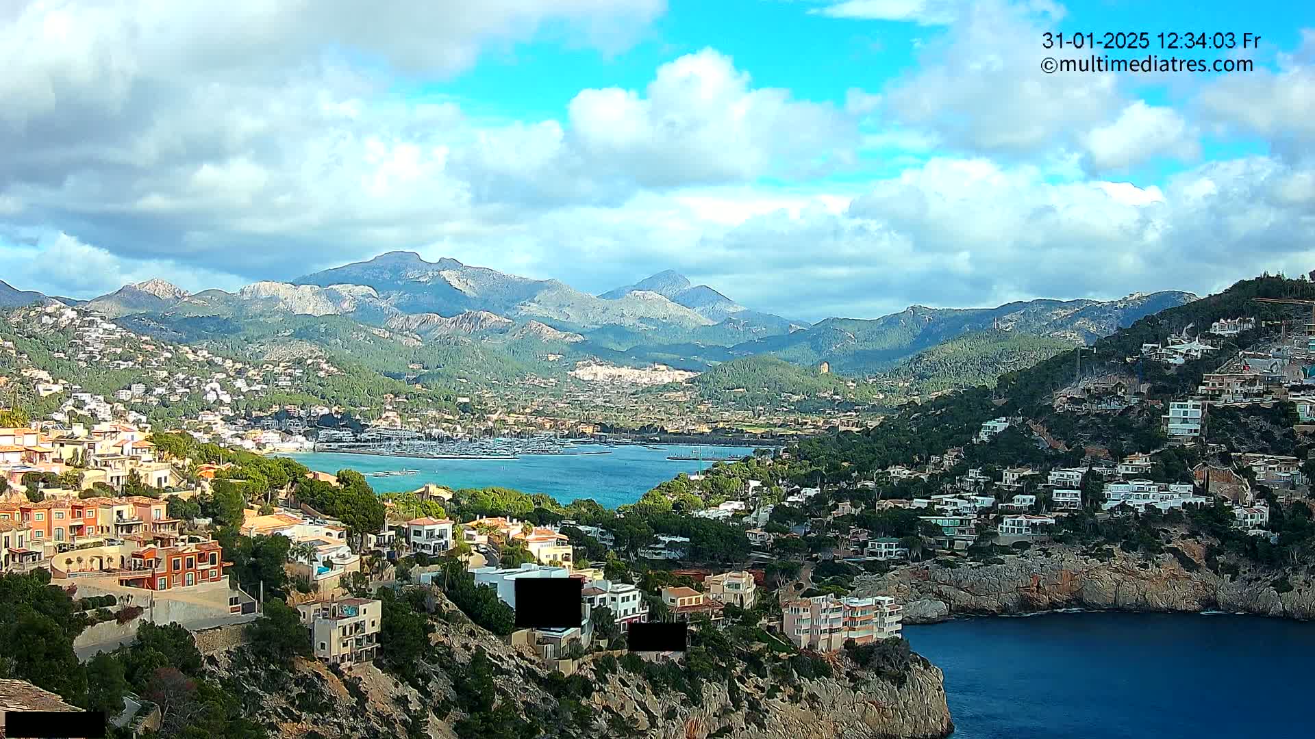 A coastal town nestled in a mountainous region under a partly cloudy sky, features a harbor filled with boats and numerous houses built into the hillsides.