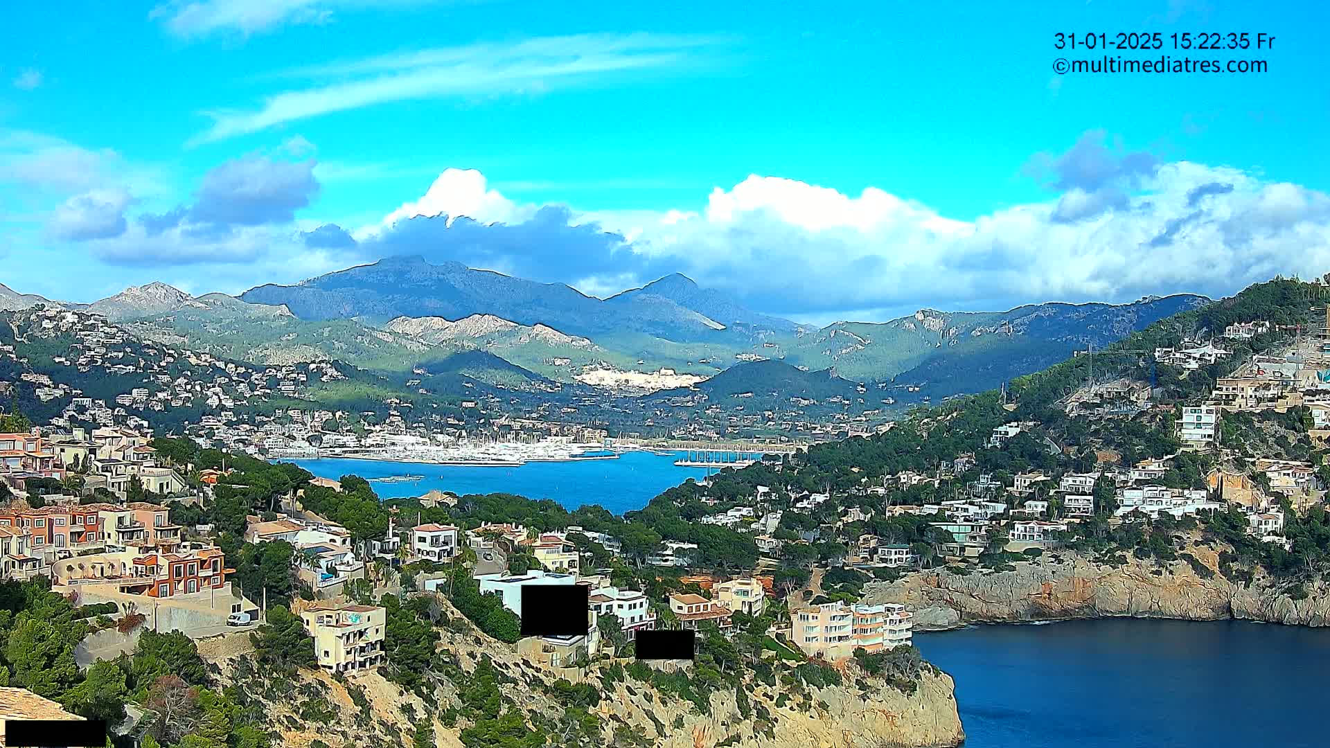A coastal town nestled among hills and mountains under a partly cloudy blue sky, with a calm body of water featuring numerous boats in a harbor.