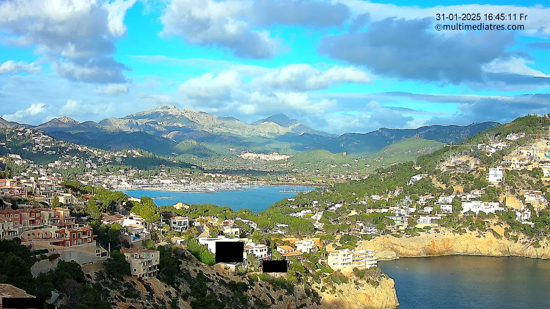 A partly cloudy day reveals a coastal town nestled in hills overlooking a calm bay and distant mountains.