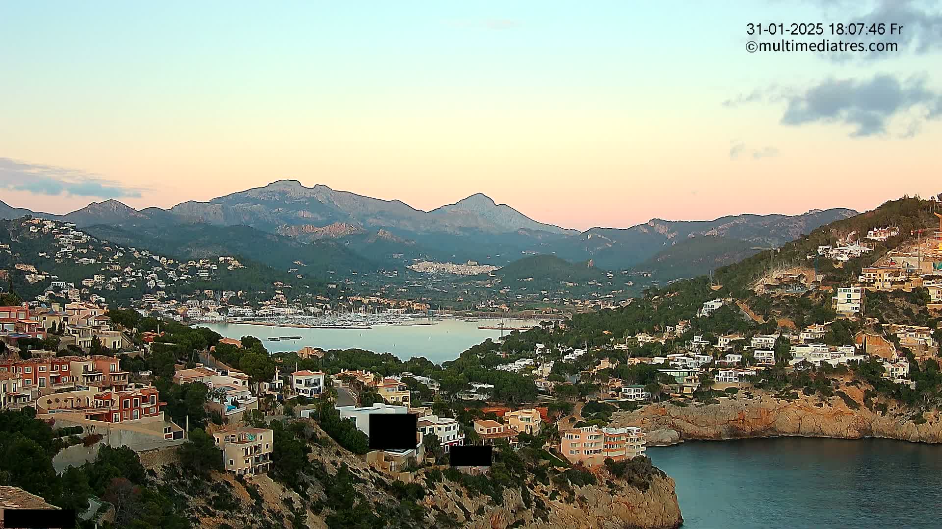 A coastal town nestled into a hillside overlooking a calm bay and a mountain range under a soft pink sunset.