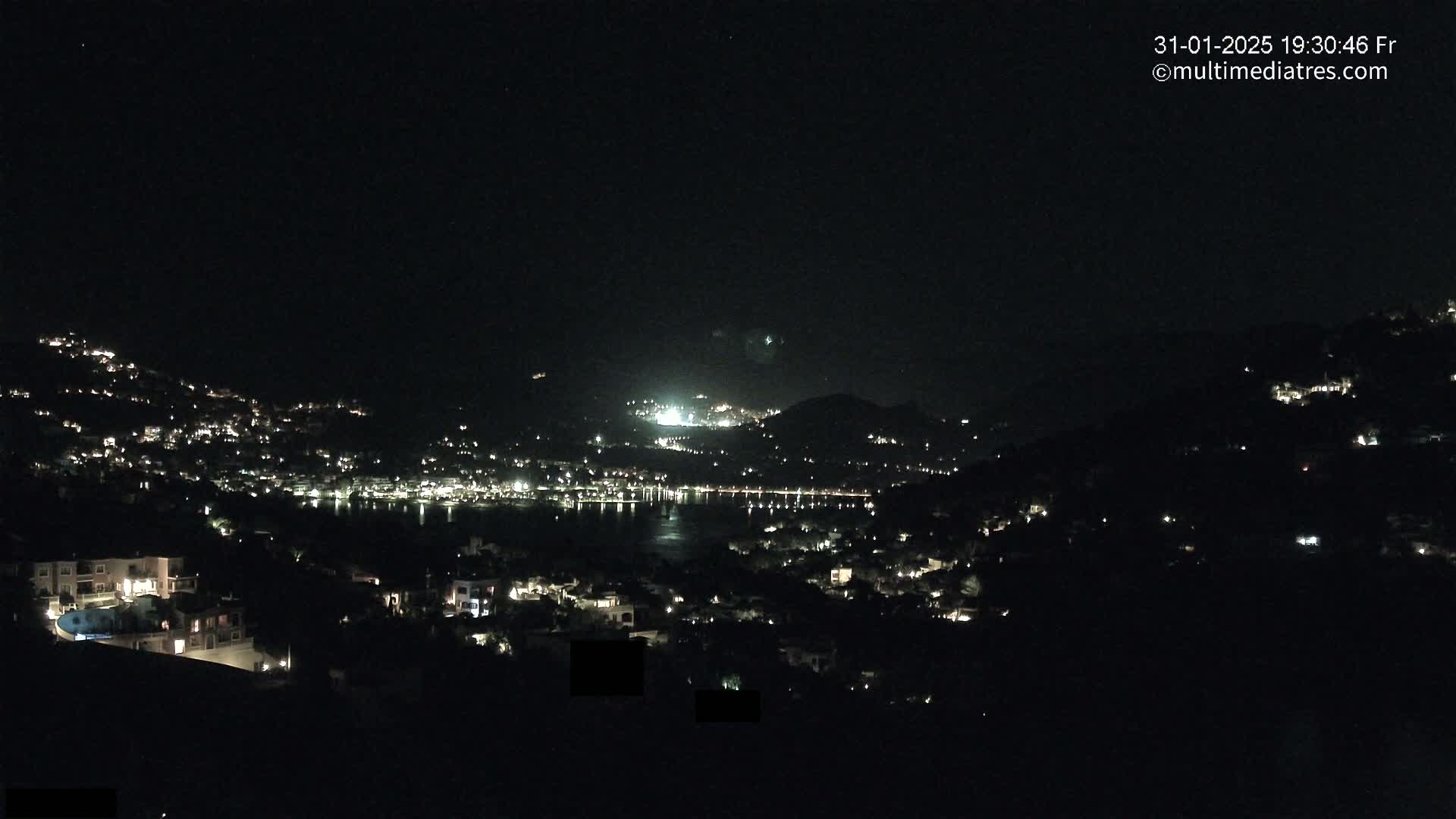 A nighttime view shows a coastal town illuminated by lights, nestled between hills and a dark body of water under a clear, starlit sky.