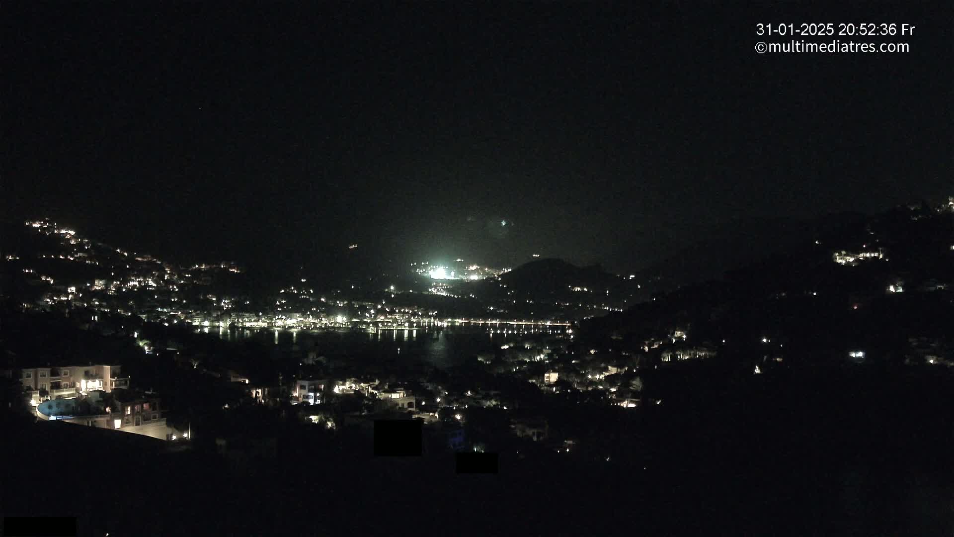 A nighttime, high-angle view shows a coastal town illuminated by scattered lights on hillsides surrounding a dark body of water.