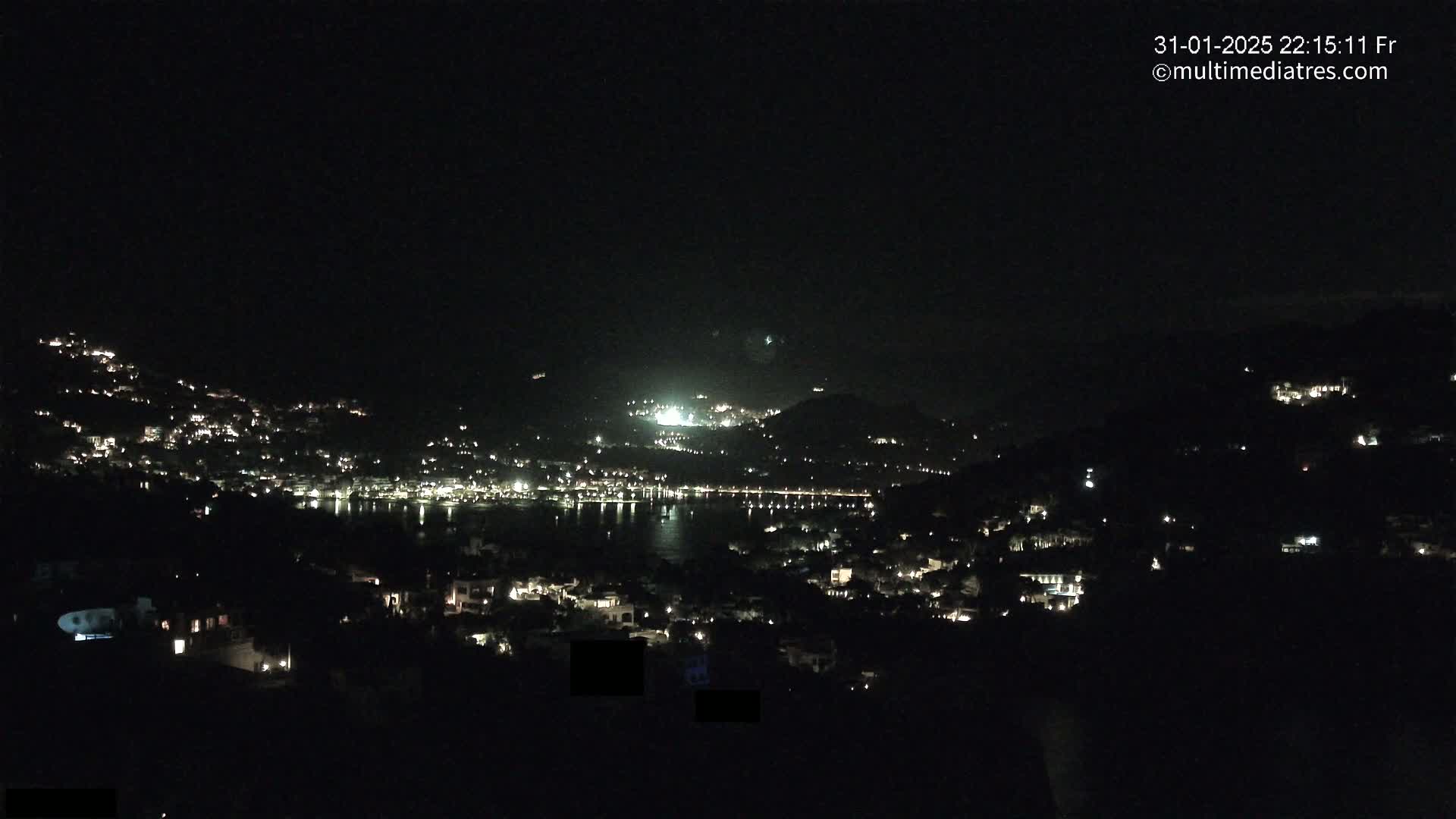 A nighttime view of a coastal town nestled on hillsides, illuminated by scattered lights, overlooking a dark body of water under a clear, dark sky.