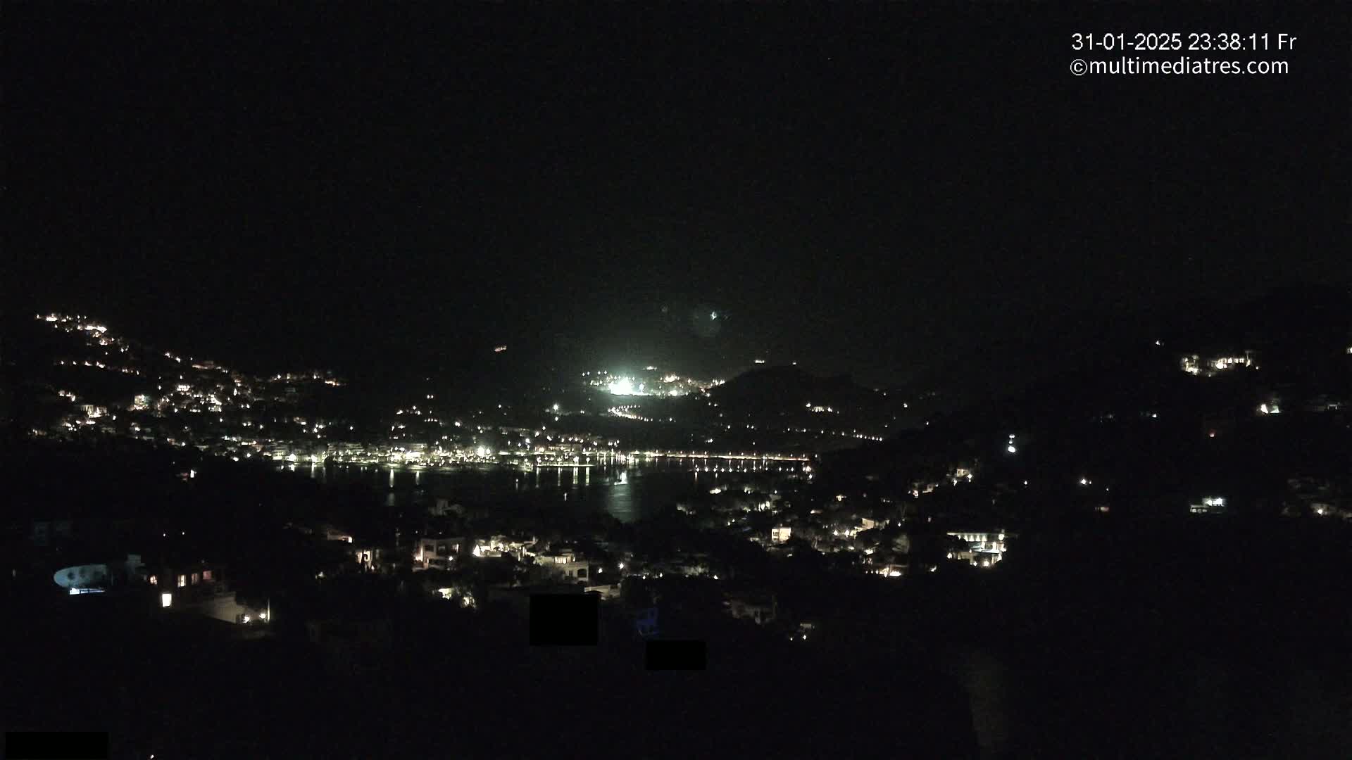 A nighttime view of a coastal town illuminated by scattered lights, nestled between hills, under a clear, dark sky.
