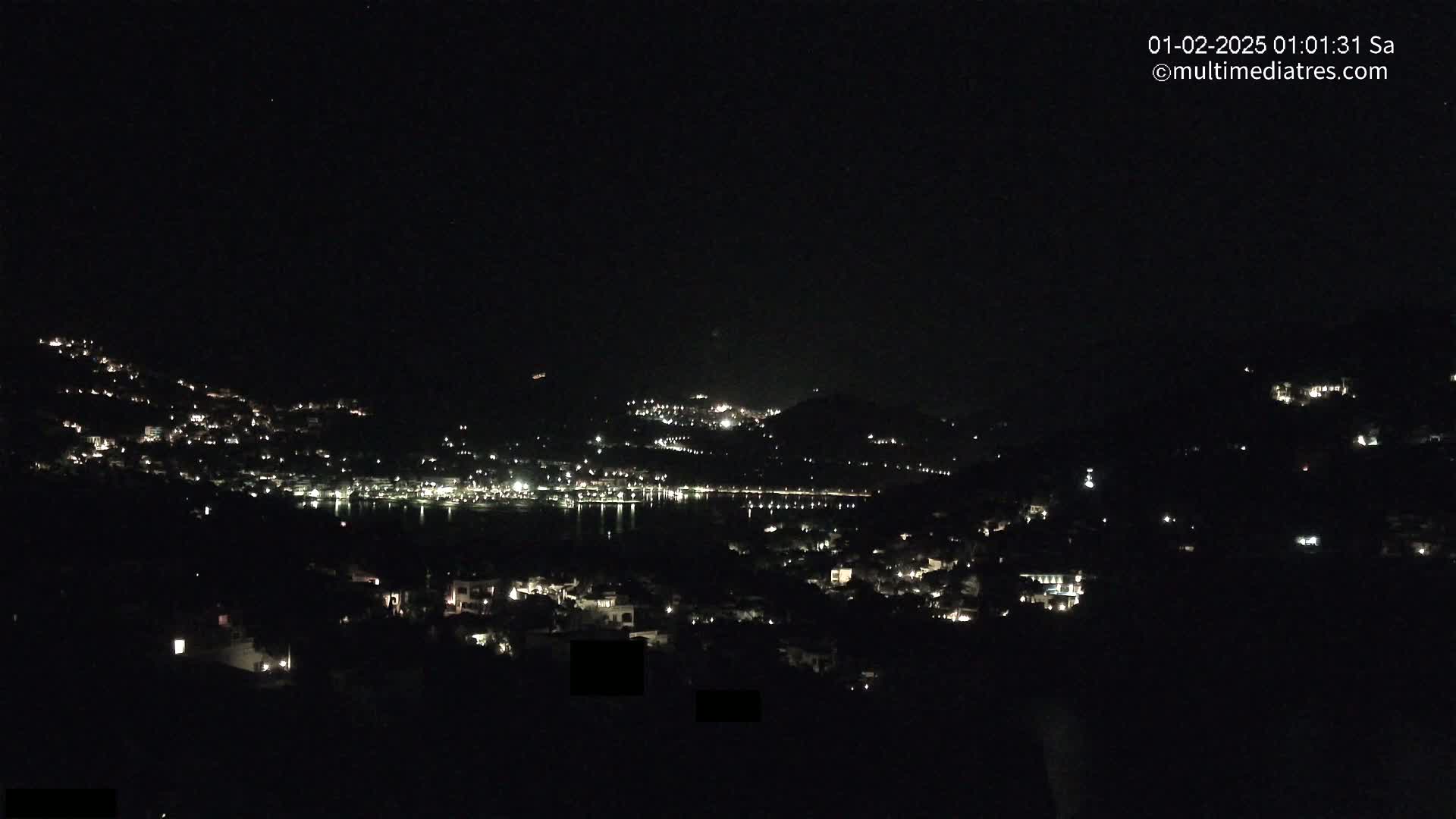 A nighttime view of a coastal town with scattered lights on hillsides surrounding a dark body of water.