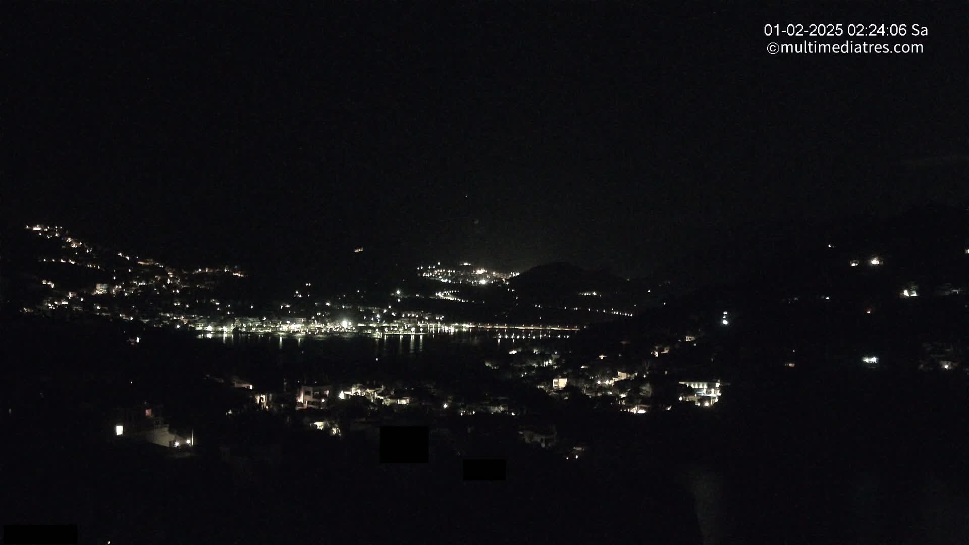 A nighttime view of a coastal town nestled between hills, with scattered lights visible across the water.