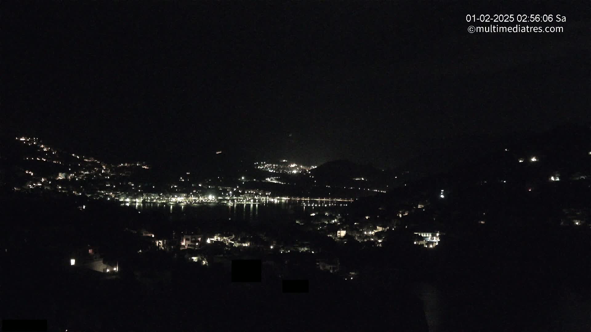A nighttime view shows a dark hillside town and a body of water, illuminated by scattered lights.