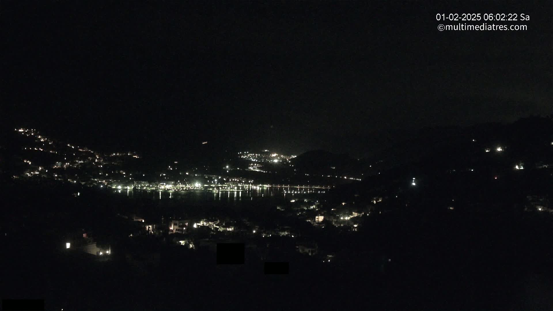 A nighttime view of a town situated on a hillside next to a body of water, with scattered lights illuminating the scene under a clear, dark sky.