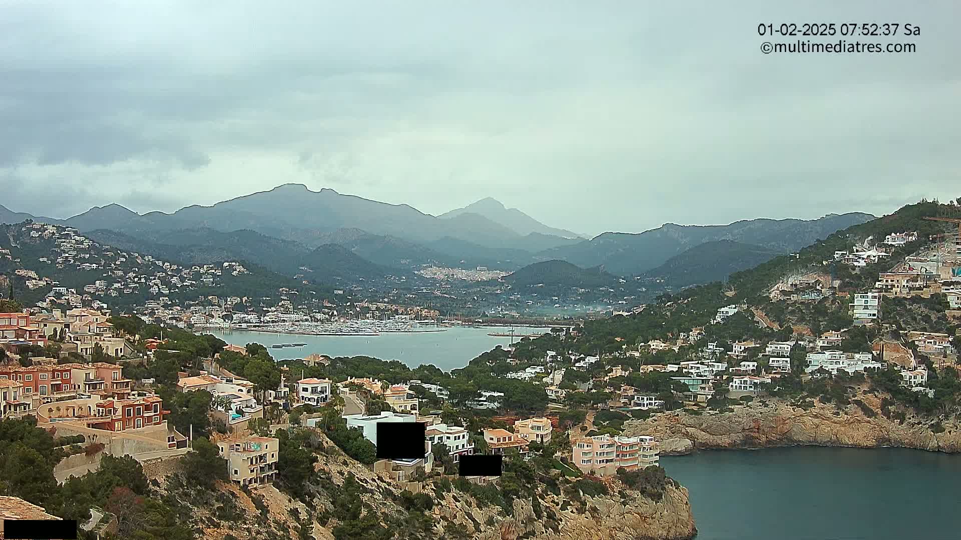 A coastal town nestled amongst hills and mountains under an overcast sky overlooks a calm bay filled with boats.