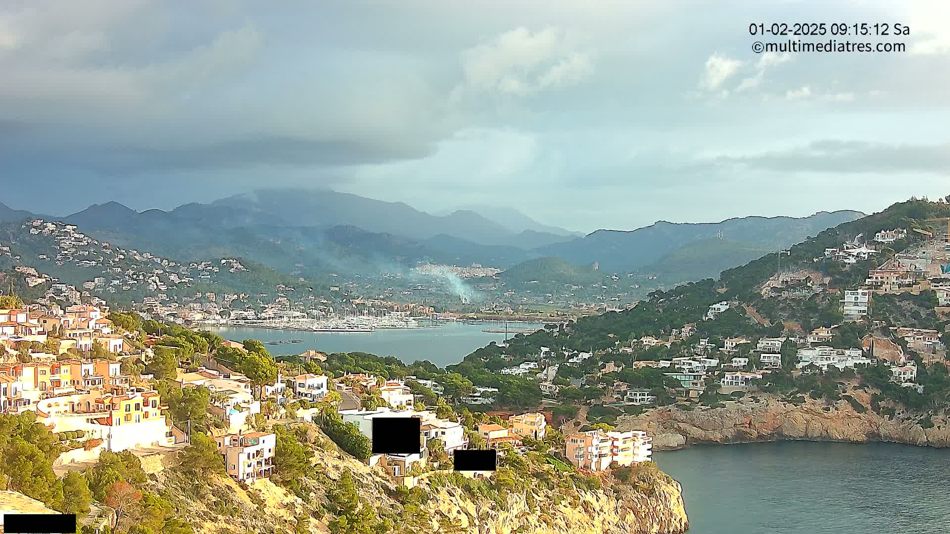 A coastal town with many houses built on a hillside overlooking a harbor filled with boats, under a partly cloudy sky.