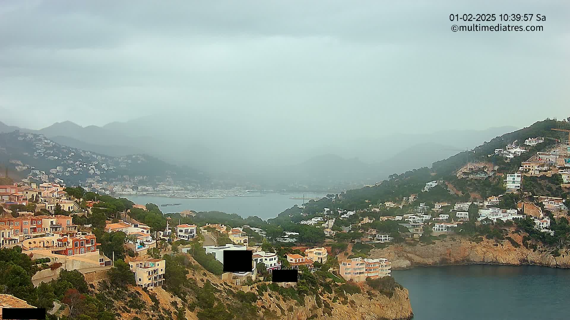 A hazy, overcast day reveals a coastal scene of numerous houses built into hillsides overlooking a calm bay with boats in the distance.
