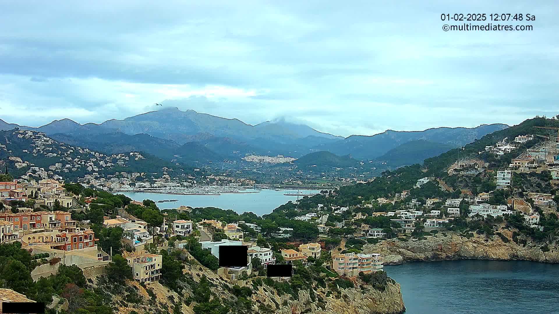 A coastal town nestled in a mountainous region under a cloudy sky overlooks a calm bay filled with boats.