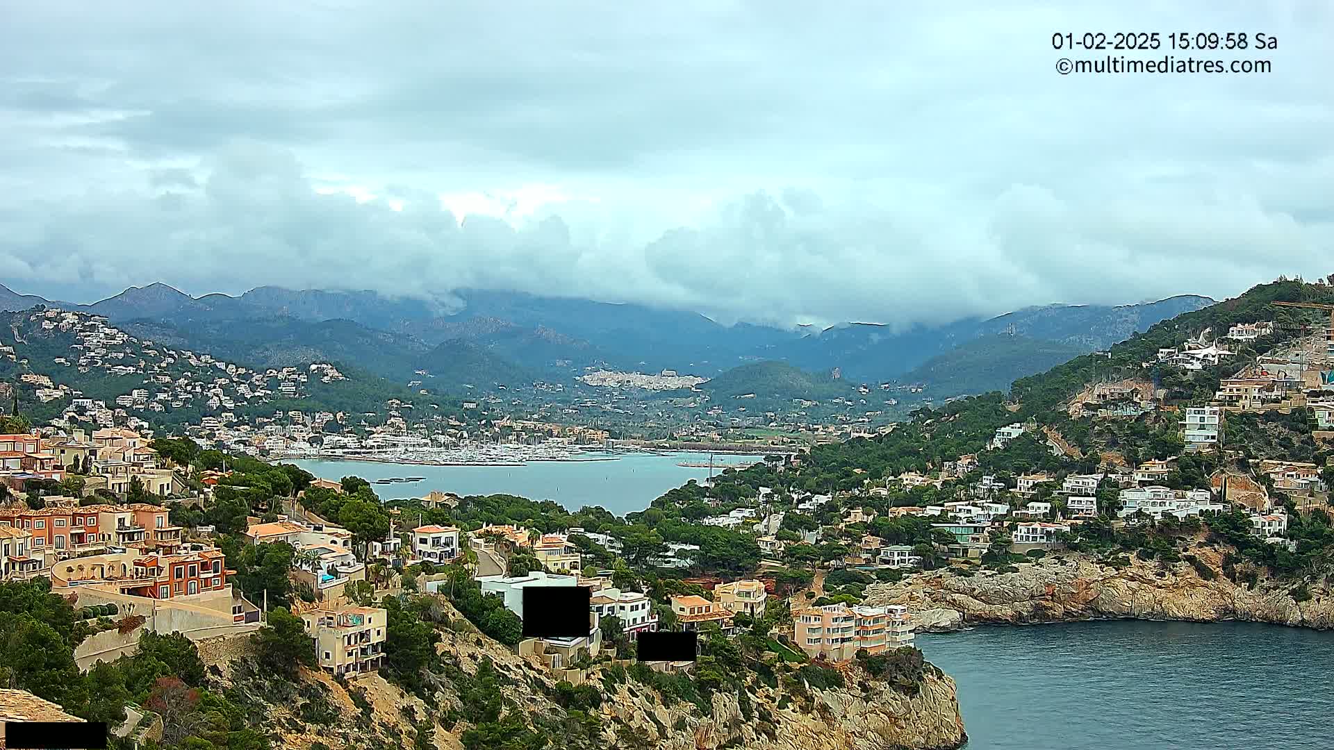 A coastal town nestled on a hillside overlooking a bay filled with boats, under a cloudy sky with mountains in the background.